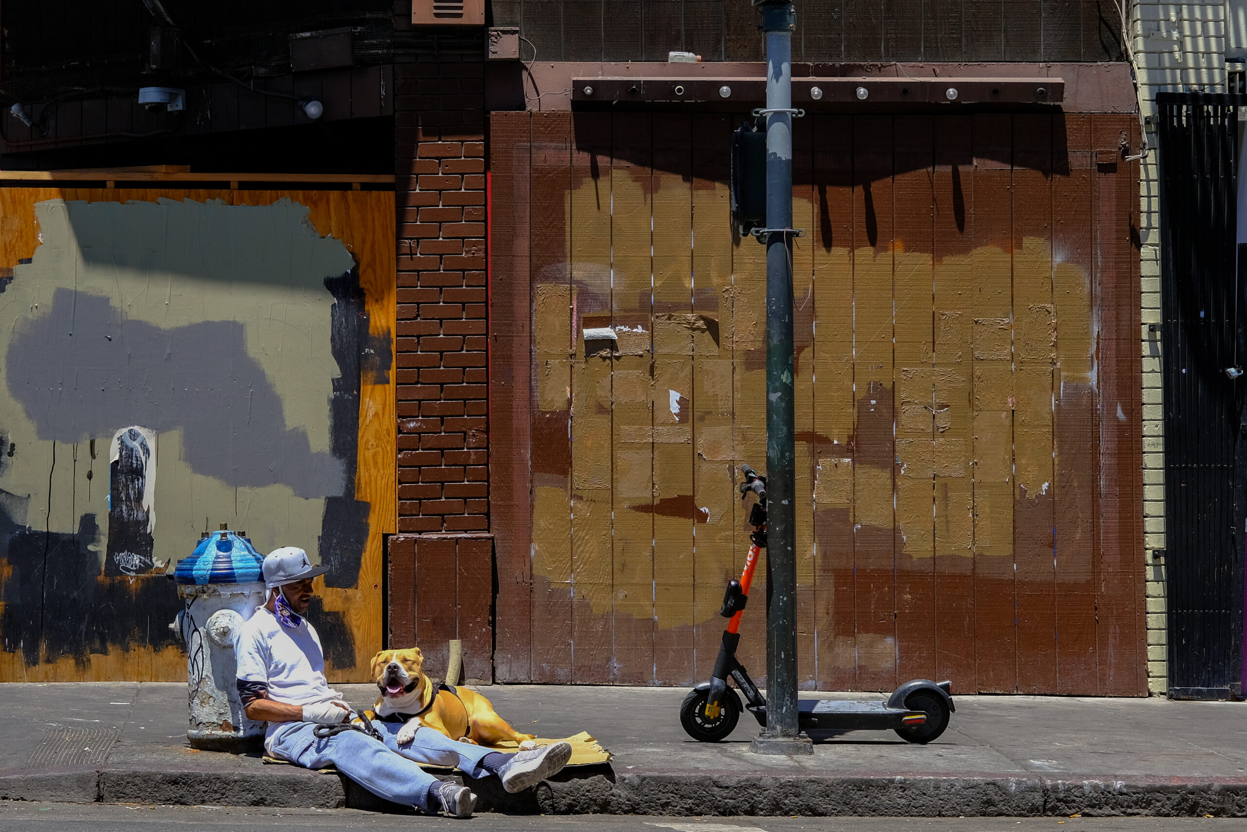 A man sits on the sidewalk next to a blue fire hydrant, accompanied by a dog. An electric scooter is nearby, and the wall behind has a weathered, painted look.