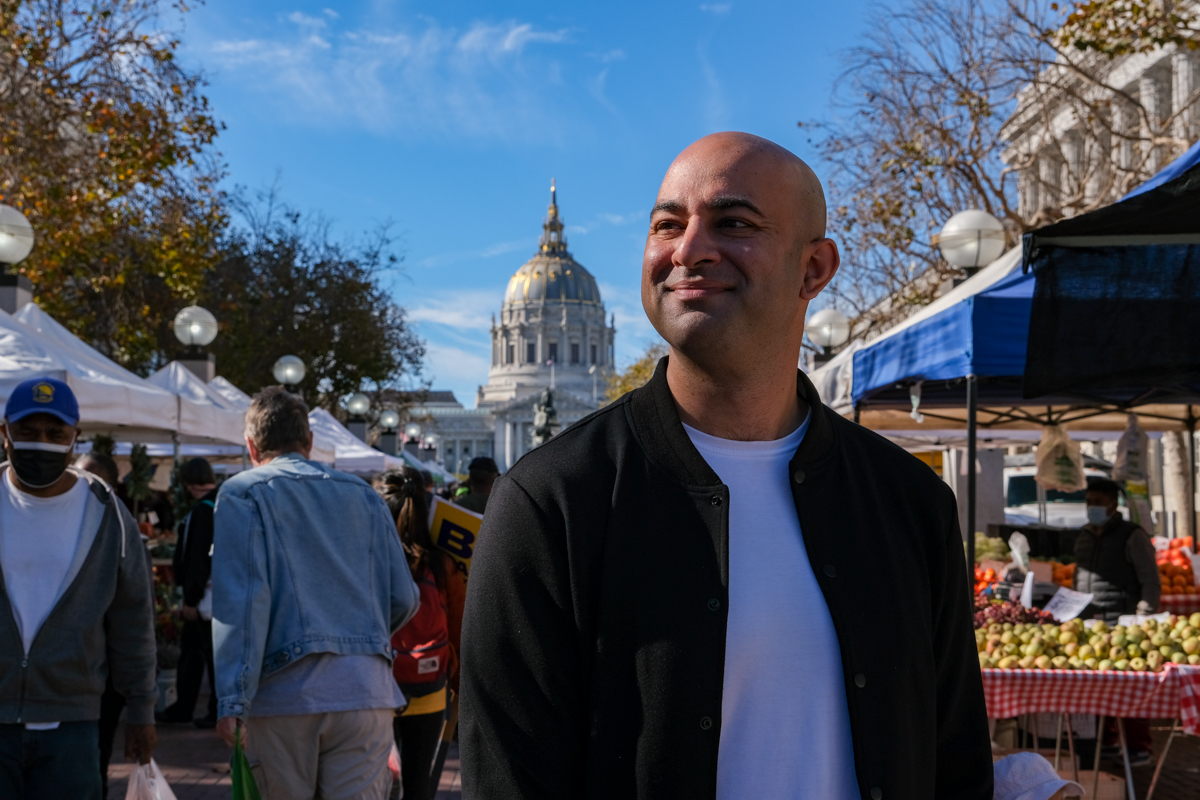Bilal Mahmood stands in a sunny outdoor market with tents, people, and San Francisco City Hall in the background.