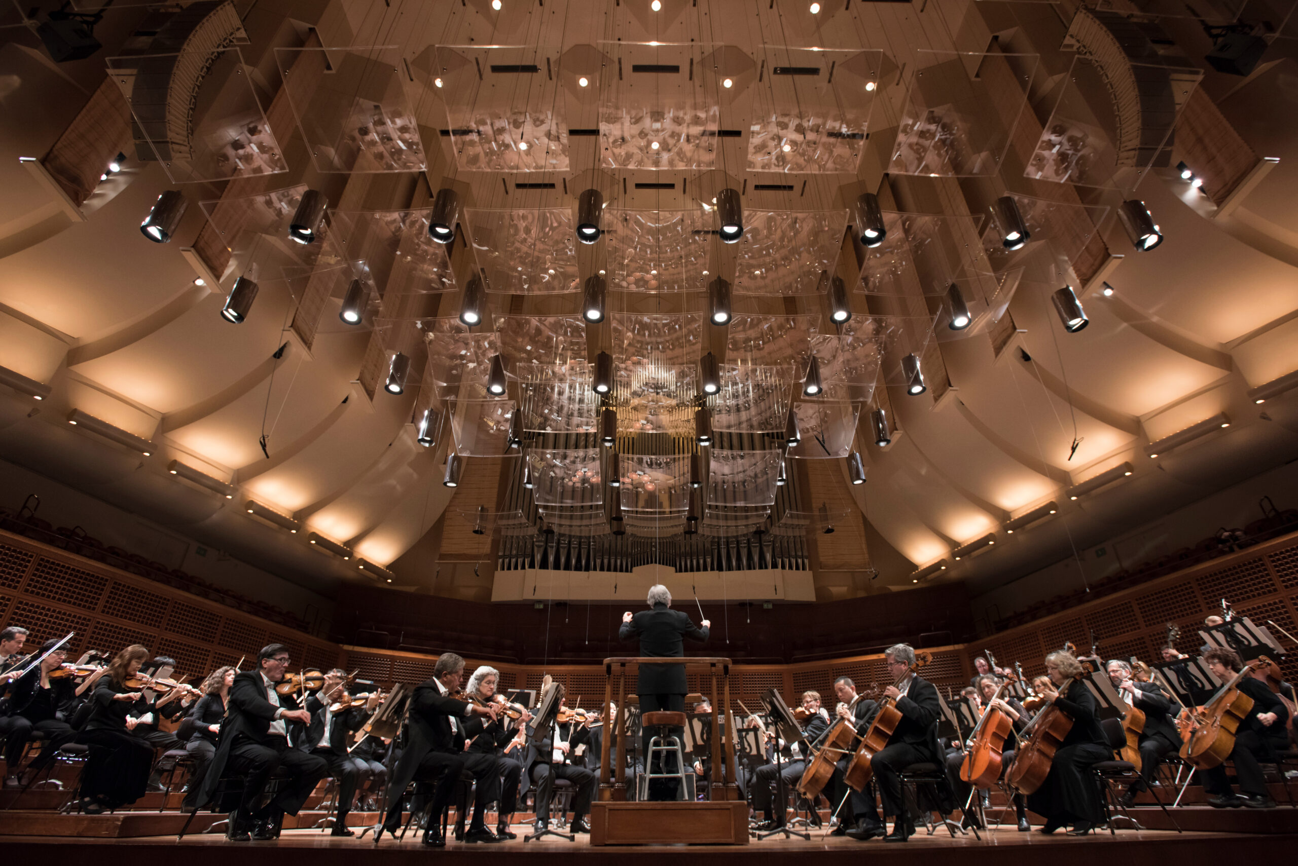 A symphony orchestra performs on stage in a grand concert hall with a conductor in front. The ceiling features numerous lights and acoustical panels.