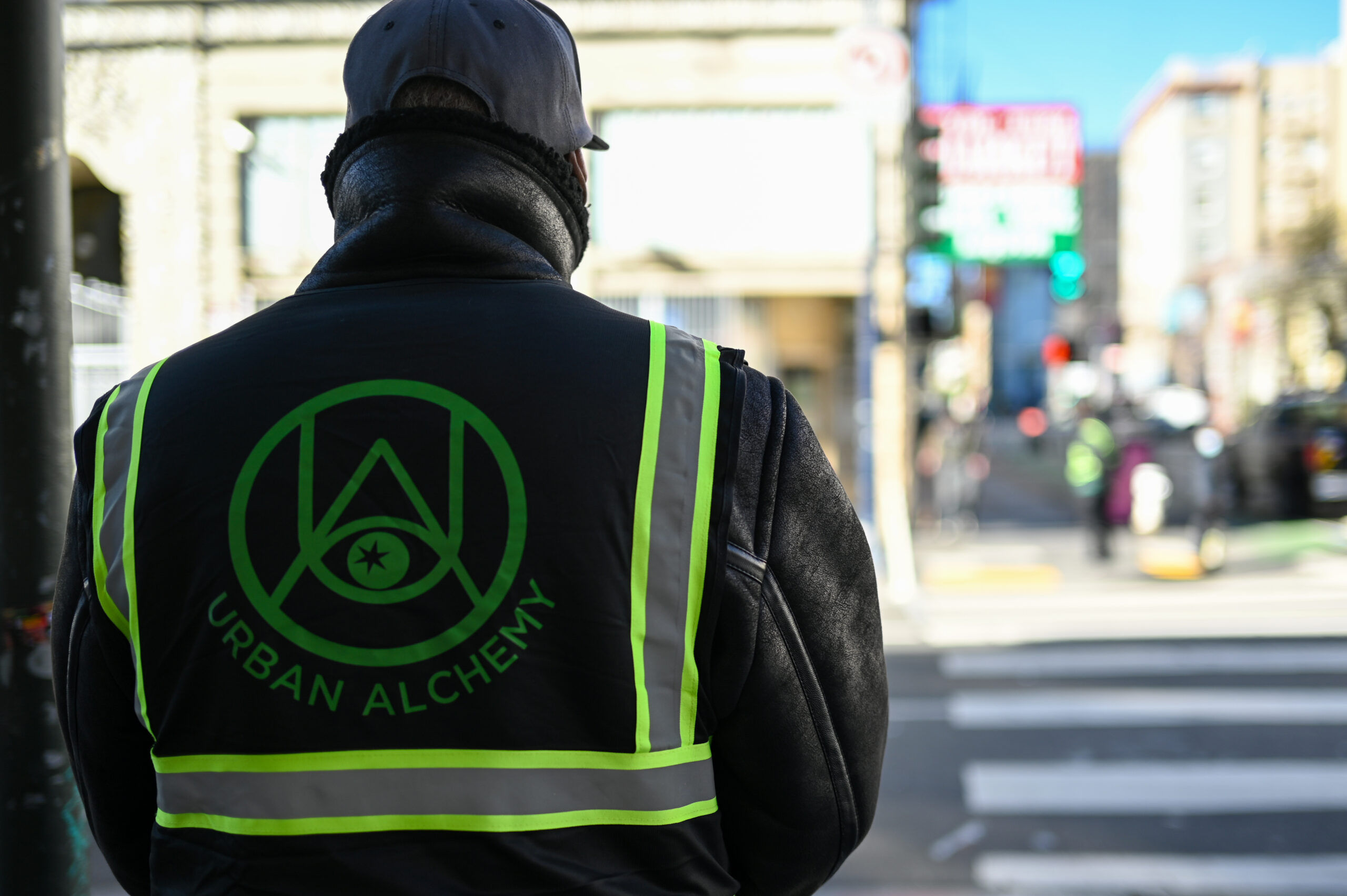 A person wearing a logo-covered vest faces away from the camera toward a city intersection.