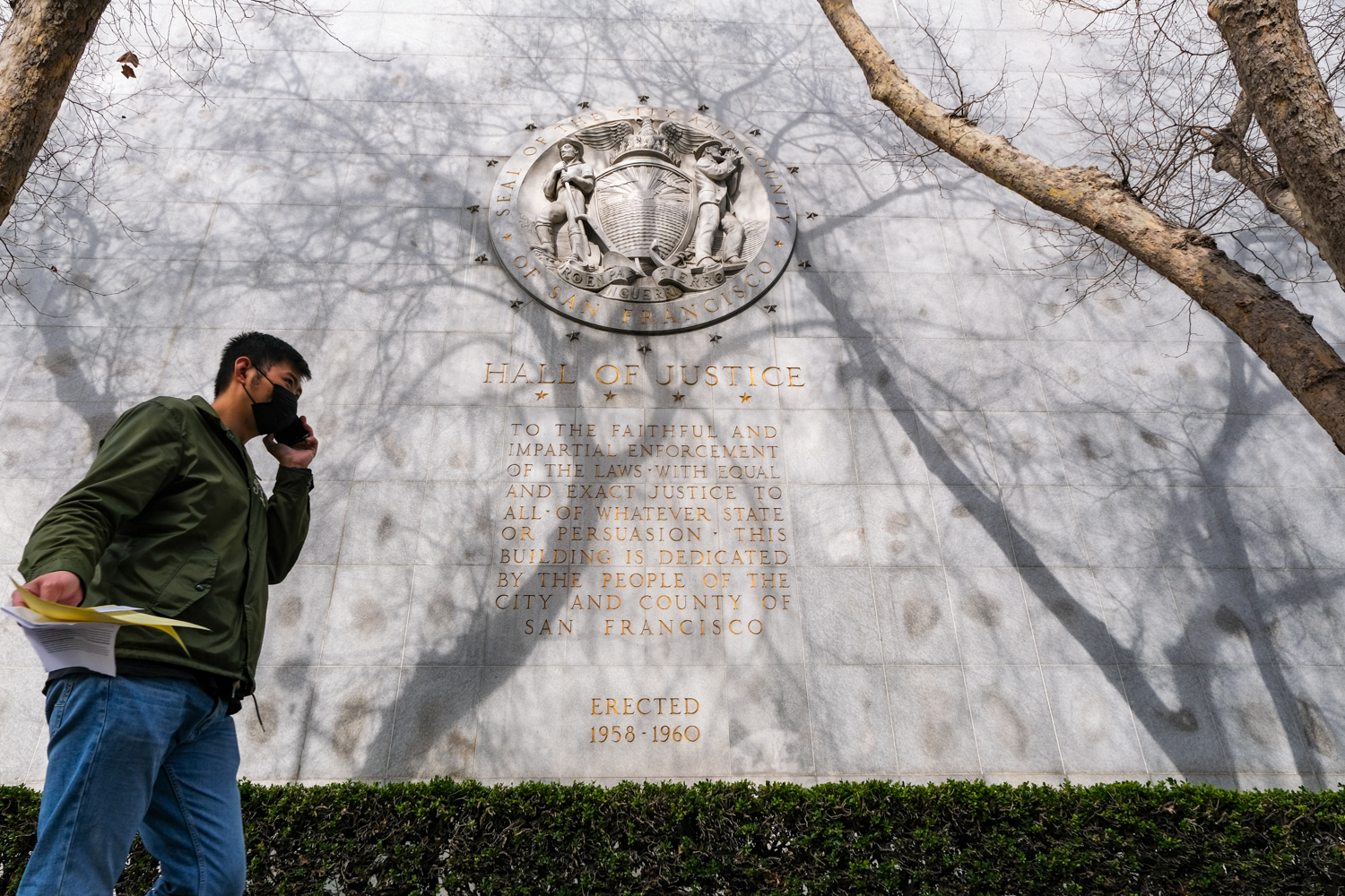 A man walks by a wall with an inscribed seal marked "Hall of Justice" and text on dedication by San Francisco's people.