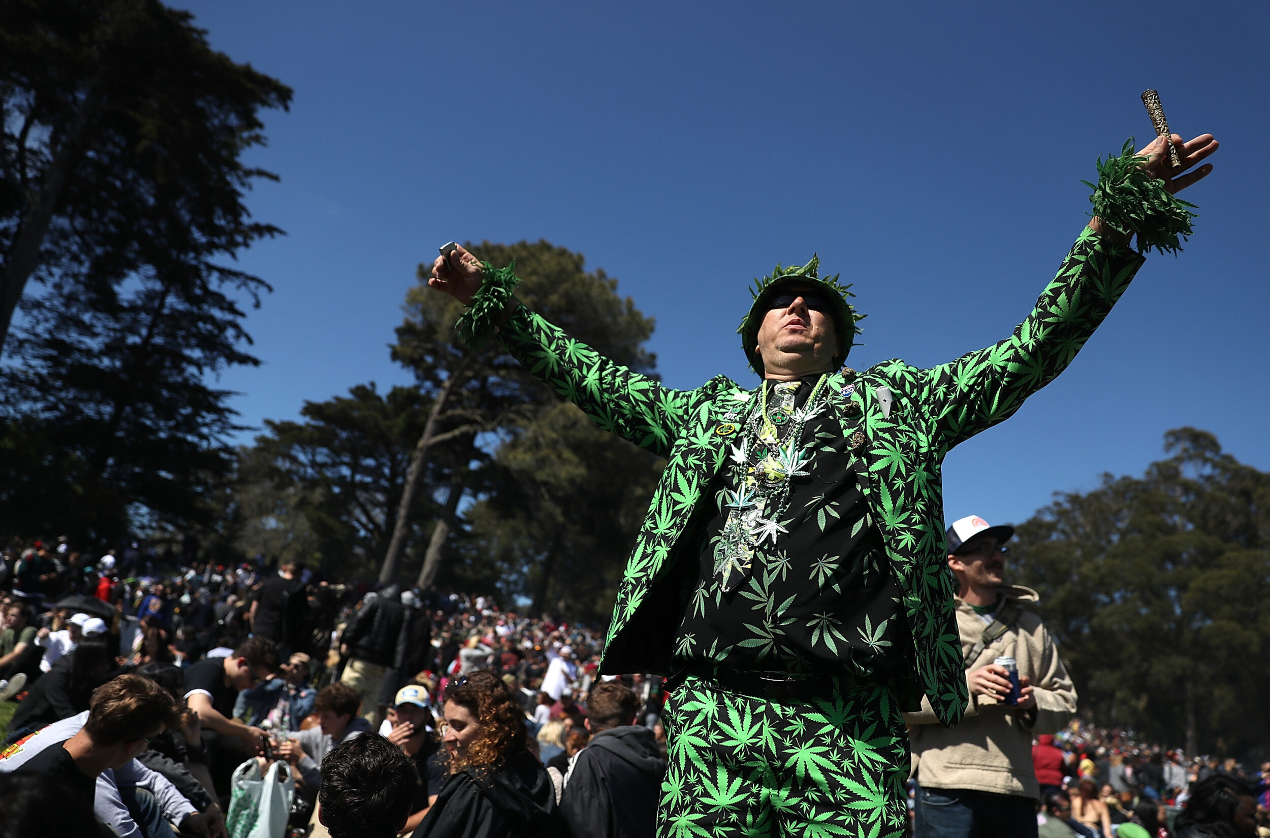 A man in a cannabis-patterned suit is standing with arms raised in a crowd under a clear sky.