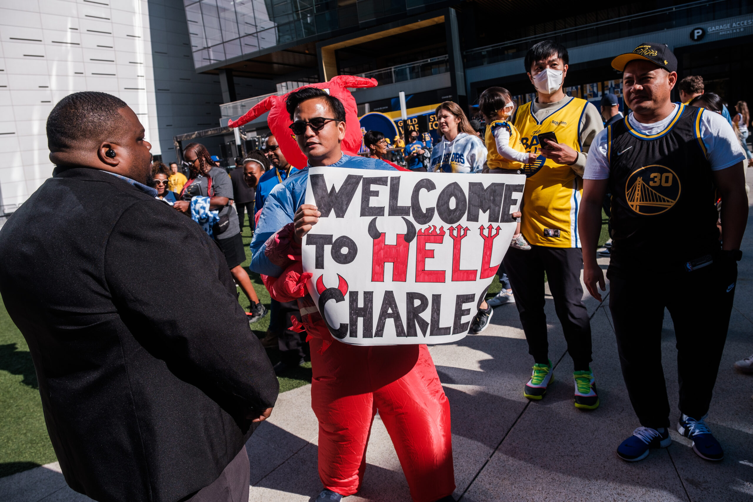 A man in red stands holding a sign saying &quot;Welcome to Hell, Charles.&quot; He's surrounded by people, including some in sports jerseys, outside a modern building.