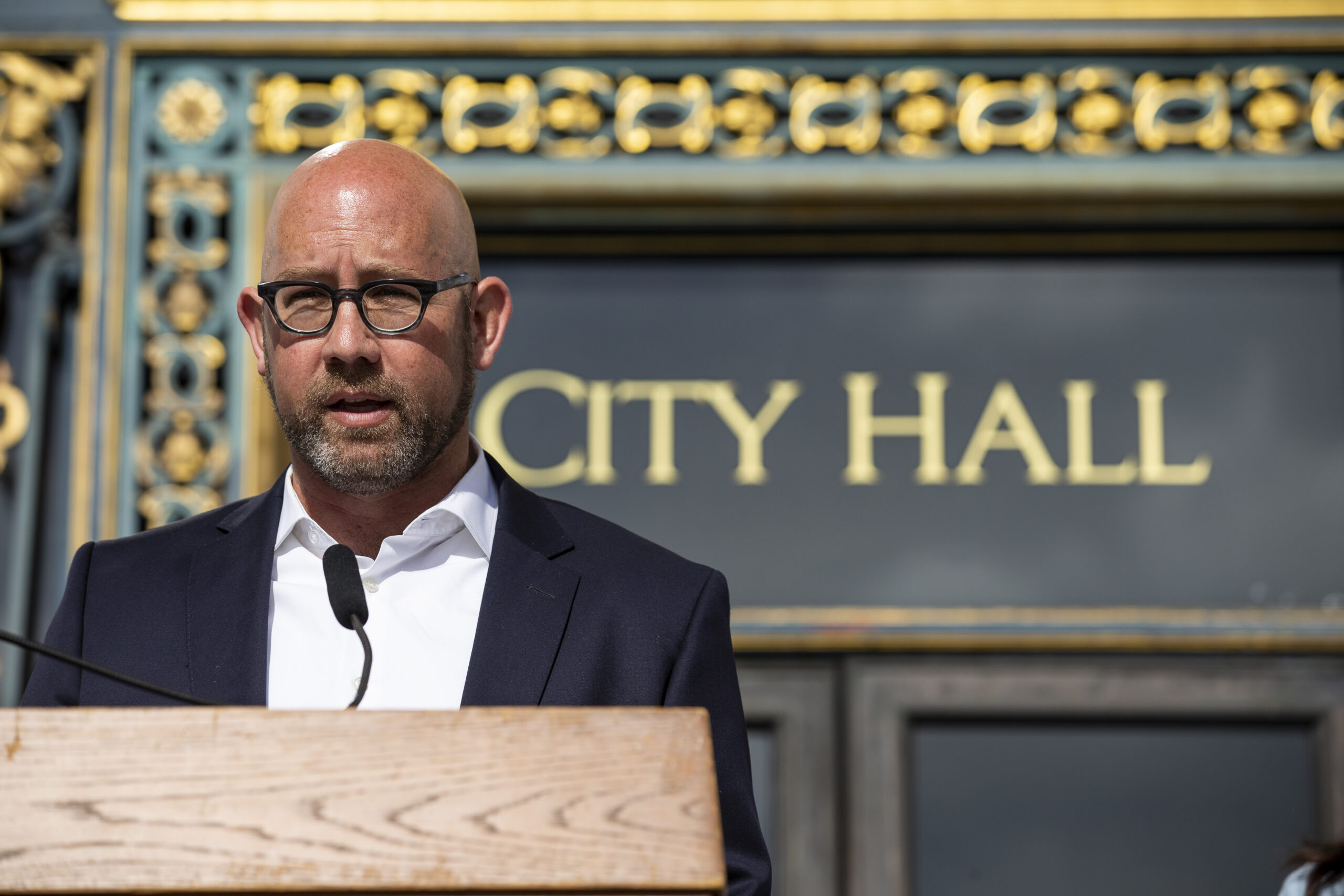 A bald man with glasses speaks at a podium marked “CITY HALL” in the background.