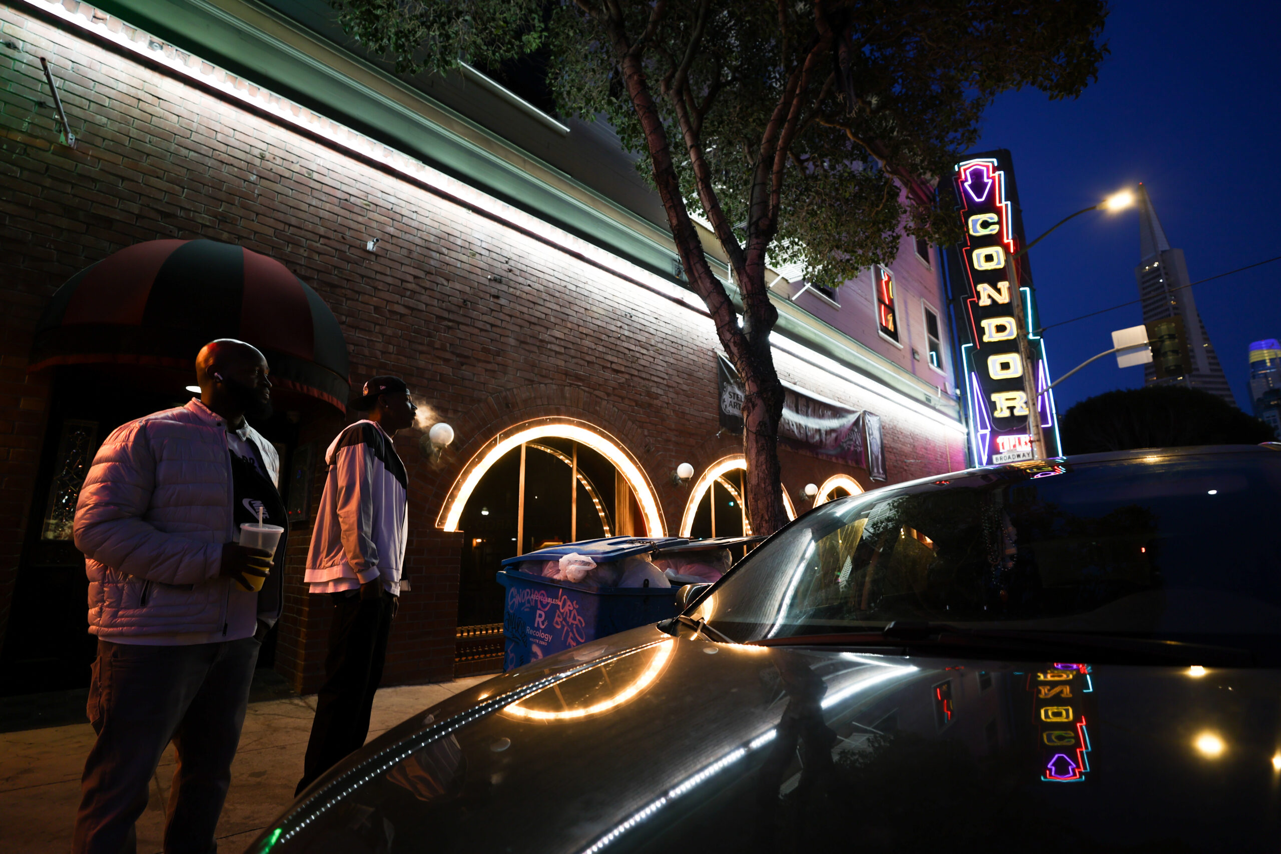 Two men stand outside a brick building at night, with a neon &quot;Condor&quot; sign and lit windows. A car is parked nearby, and a tree is visible overhead.