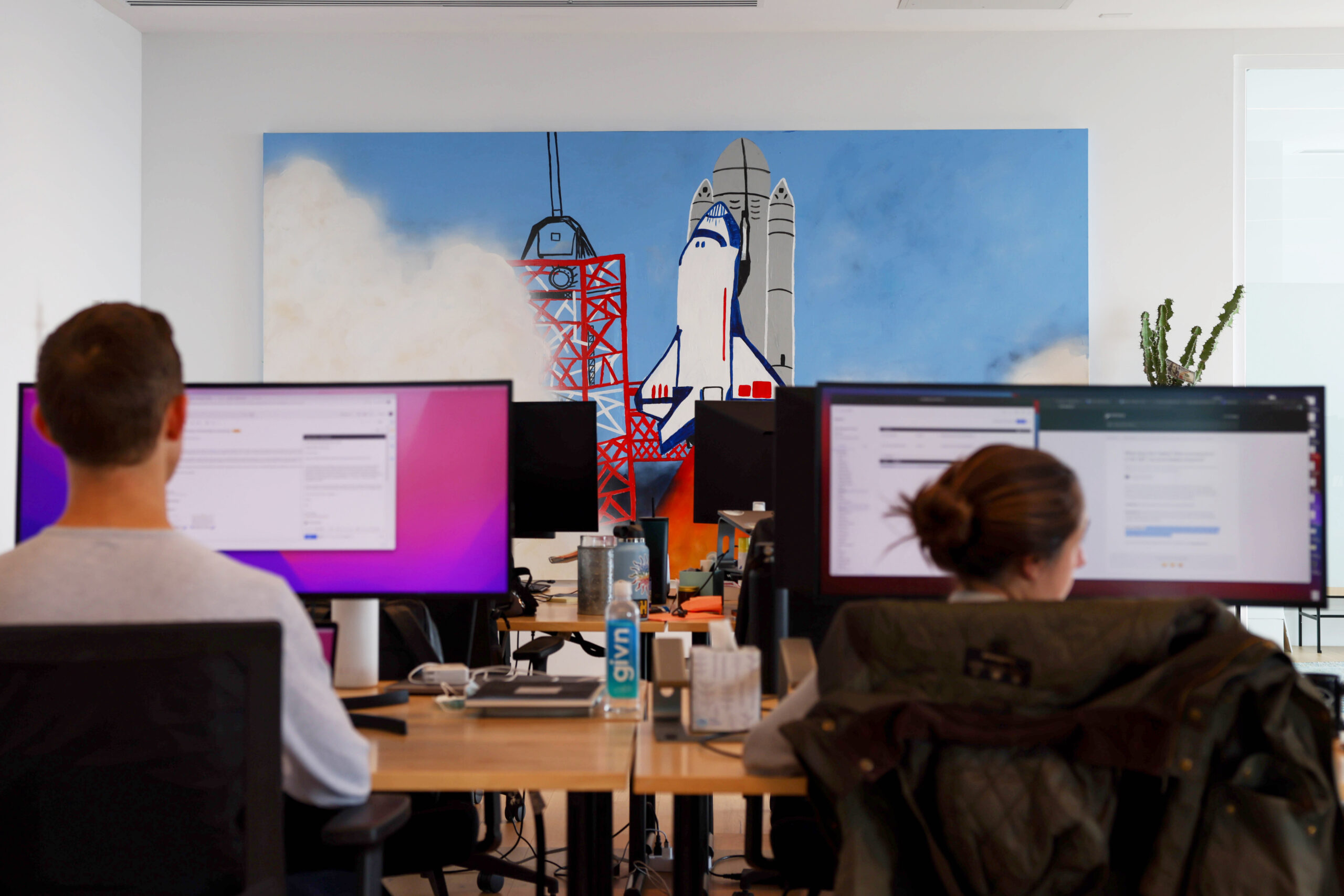 A photo of two people working at their computers in a startup office with a space shuttle painting in the background.