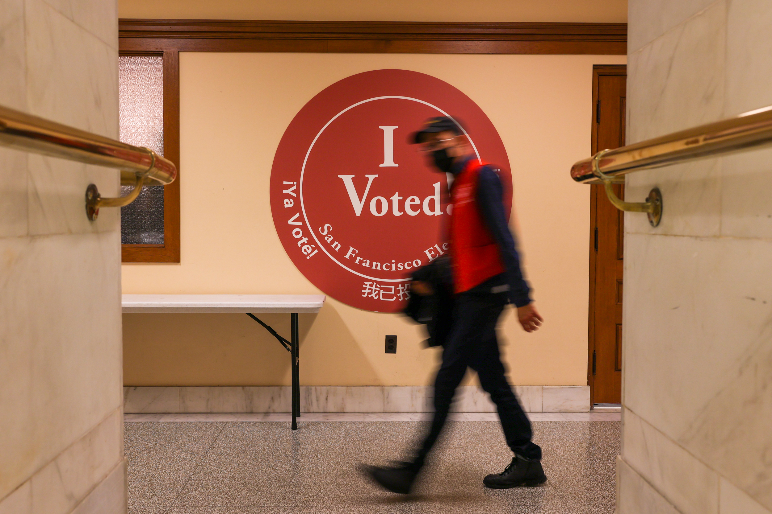 A blurred person walks by a large "I Voted" sticker sign with text in multiple languages, indicating a polling station.