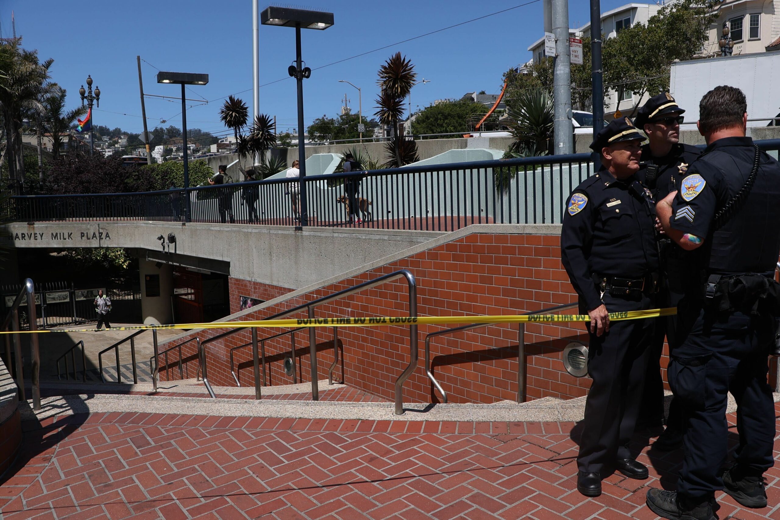 Two police officers stand by a taped-off urban area near a plaza entrance with palm trees and a clear sky.