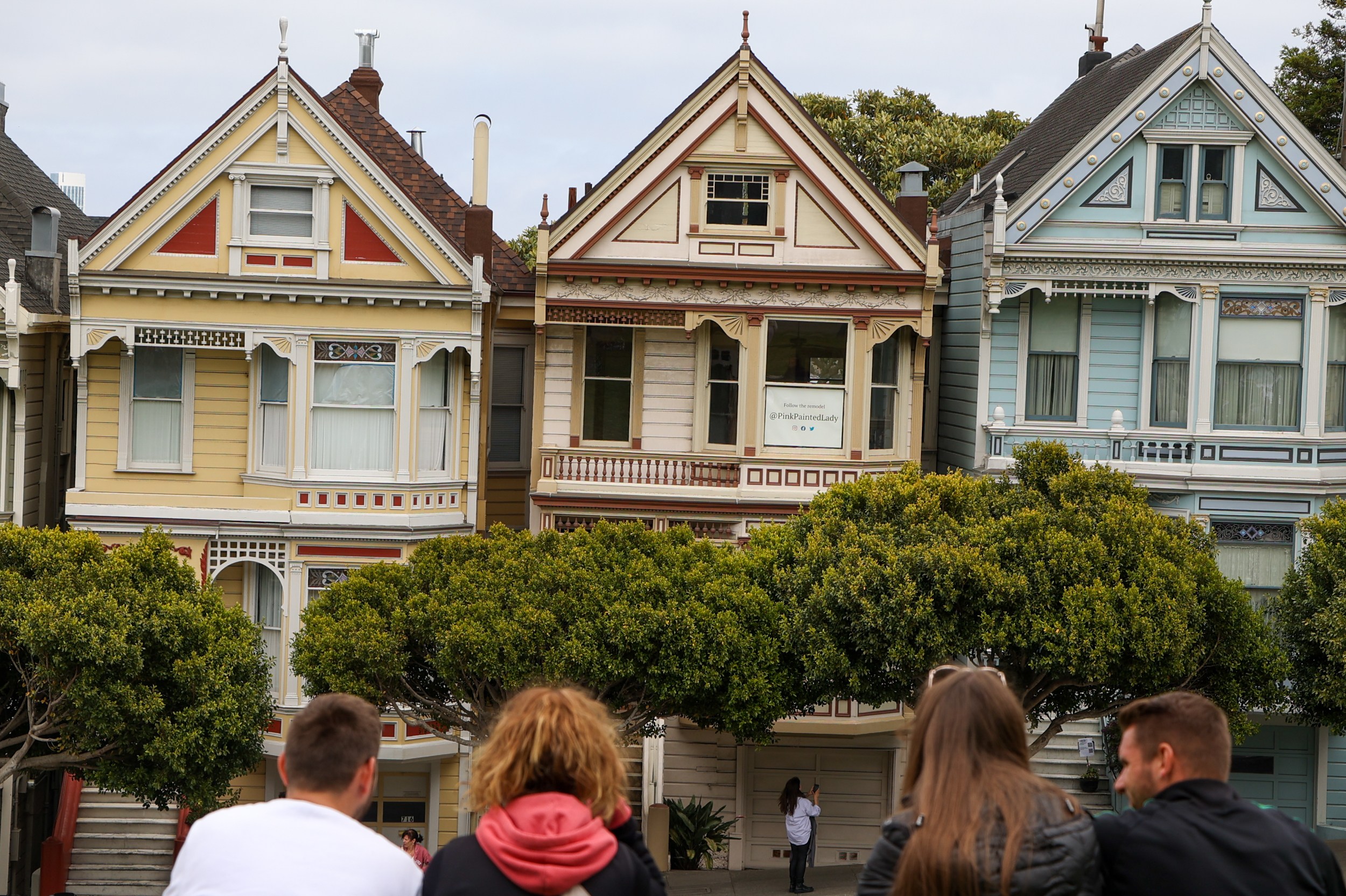 Three ornate Victorian-style houses, each with distinct colors and architectural details, are in view. In the foreground, four people are sitting and observing.