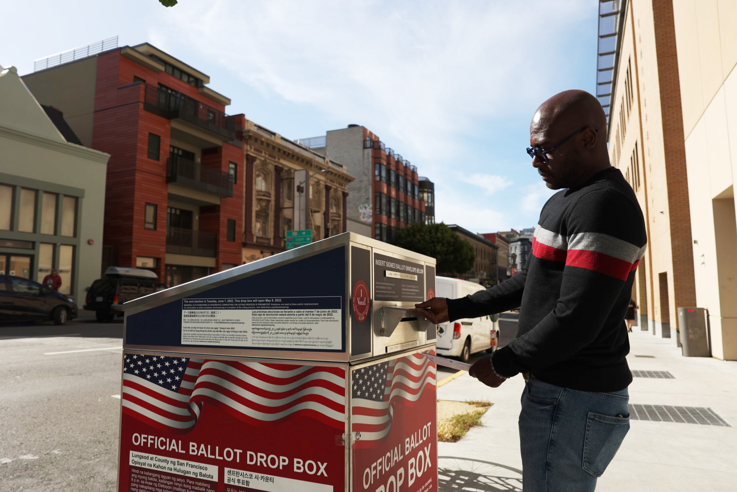 A man wearing sunglasses prepares to insert his ballot into a drop box on a sunny San Francisco street.