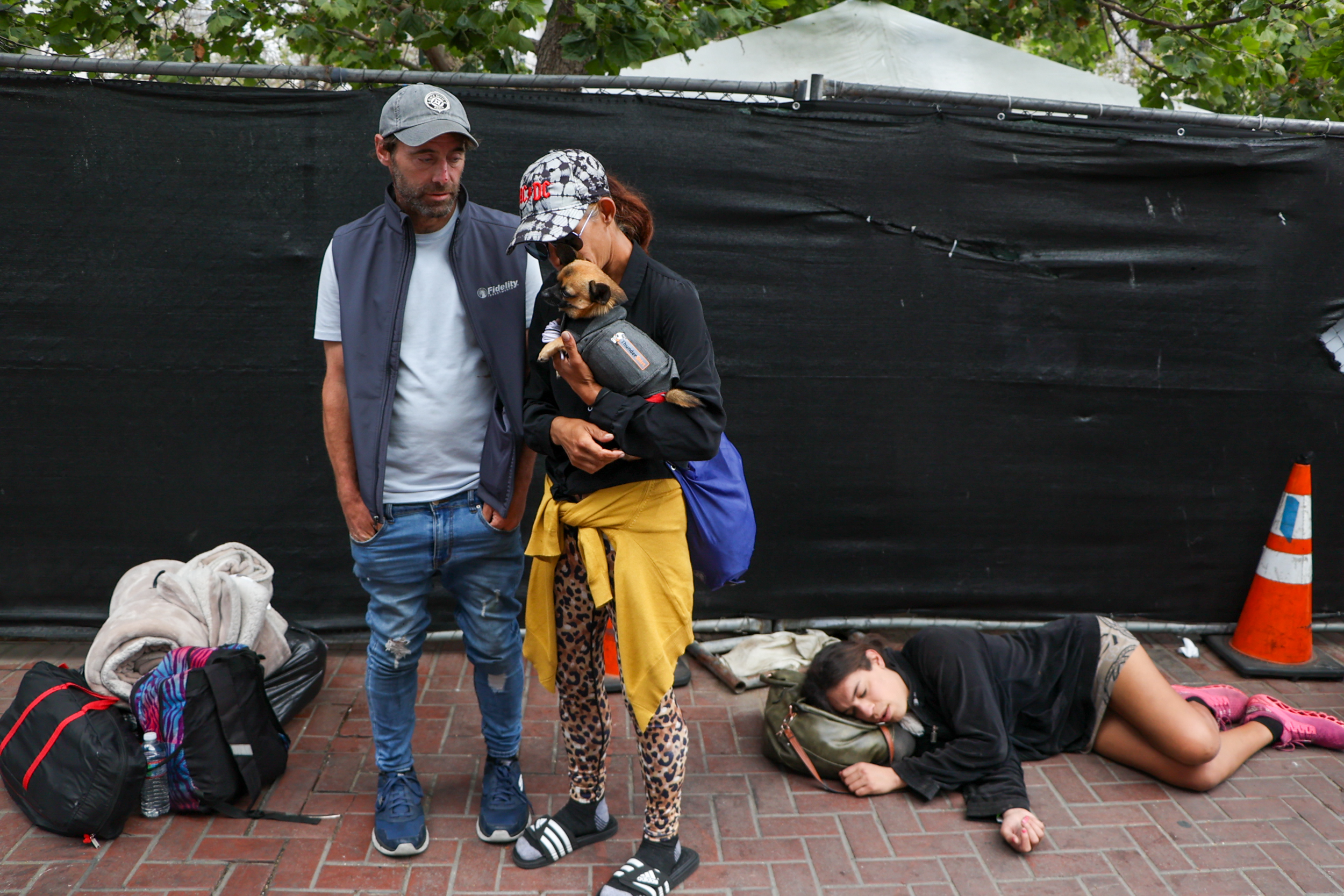Two adults stand with a dog; a woman sleeps on the pavement nearby, with bags and a cone visible.