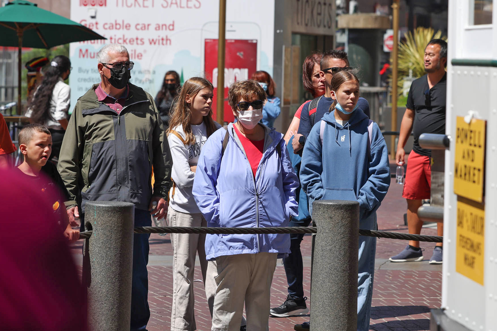 A group of people are seen, some are wearing masks and rain coats on a city street. Some of the people are not wearing masks.