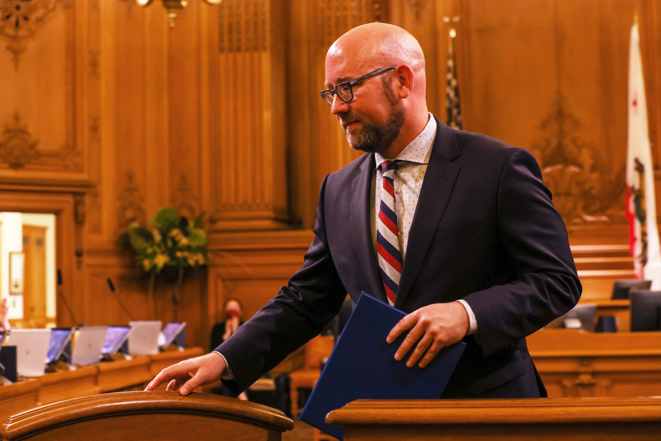A man in black-rimmed glasses and a dark blue suit walks through a gate inside City Hall's Board of Supervisors' chambers.