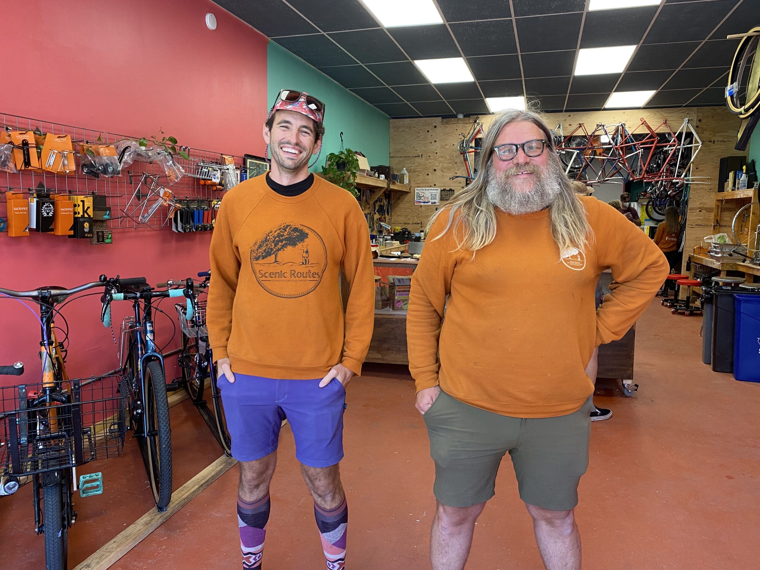 two white men in orange sweatshirts stand with their hands in their pockets inside a bike shop