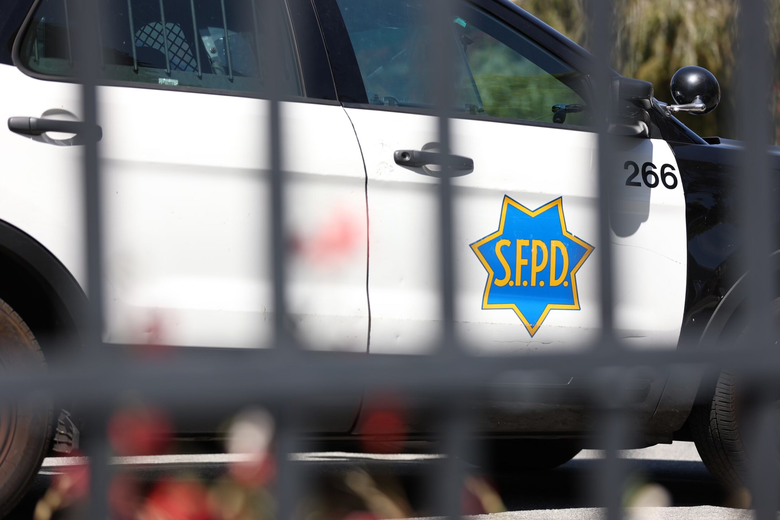 A police car with "S.F.P.D." badge, viewed through a blurred fence.