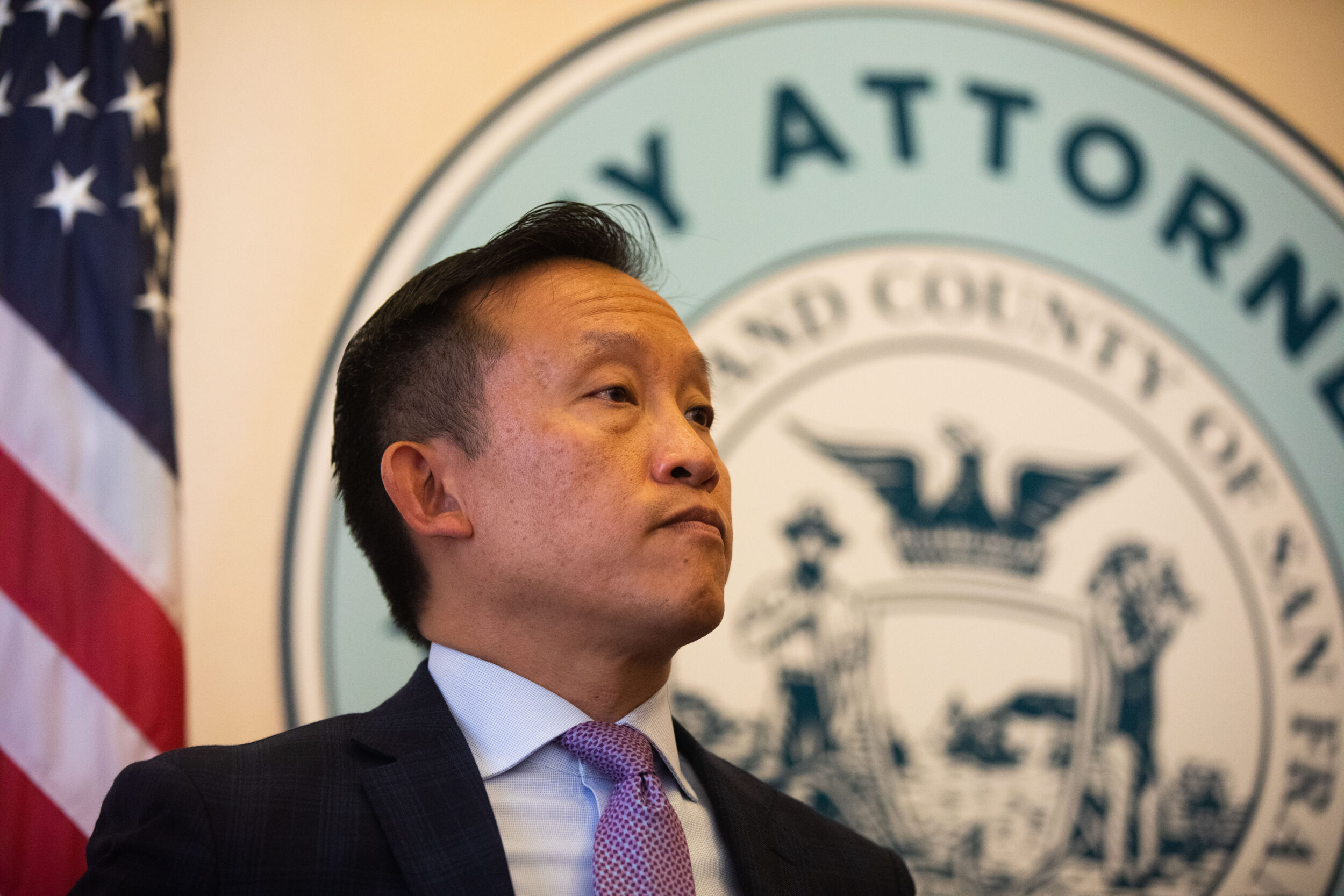 City Attorney David Chiu, wearing a suit and tie, looks on in front of the City Attorney's official seal and an American flag.