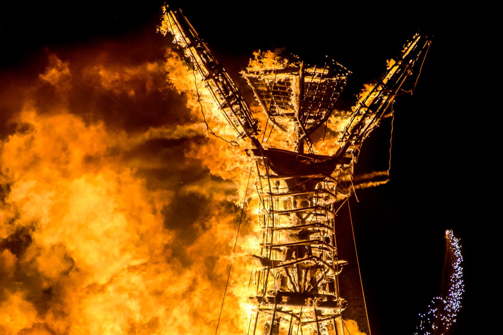 A large wooden structure resembling a human figure is engulfed in bright orange flames against a dark sky, creating a dramatic, fiery spectacle.