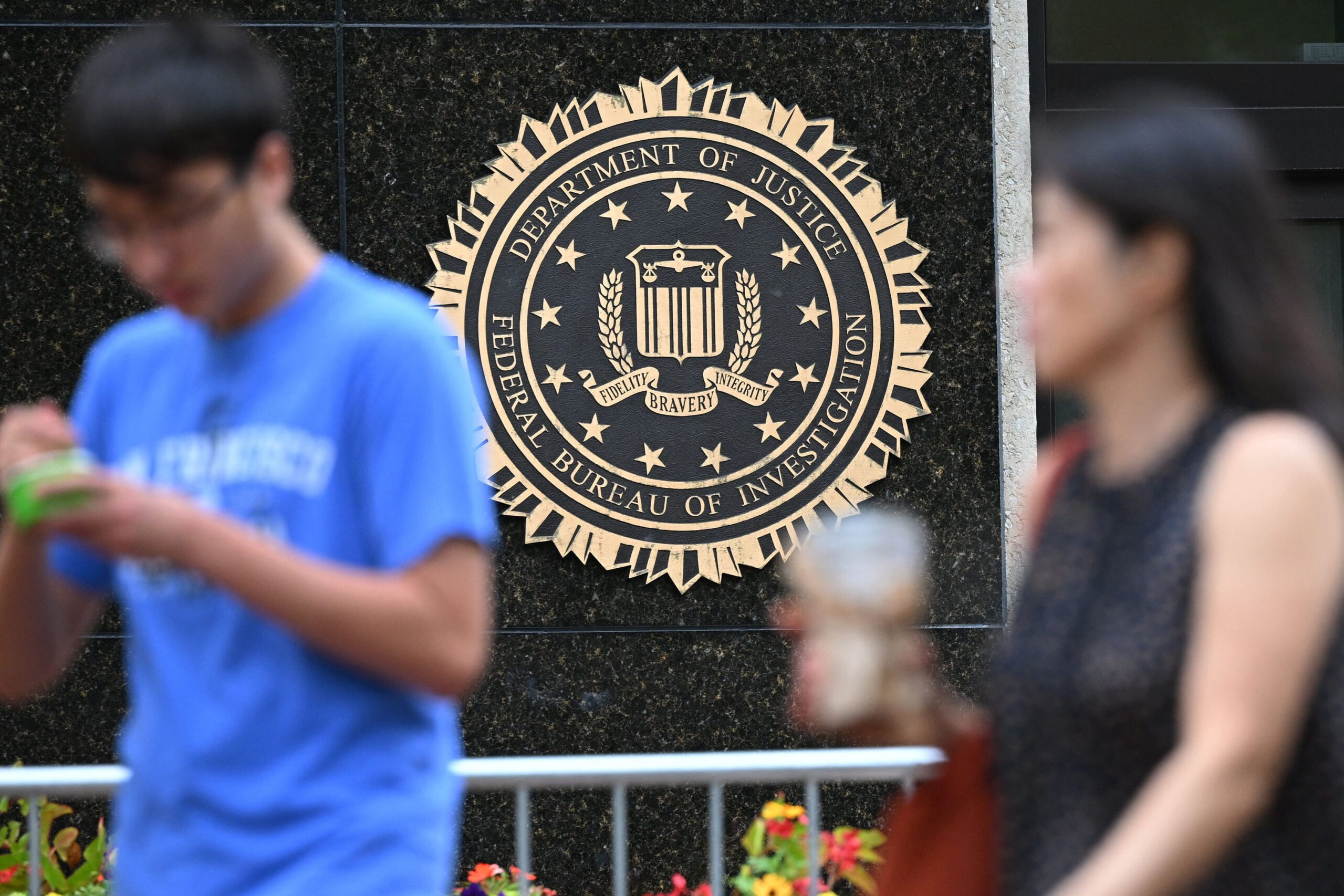 The image shows the FBI seal on a building facade, featuring the Department of Justice emblem. Two blurred figures are in the foreground, one holding a drink.