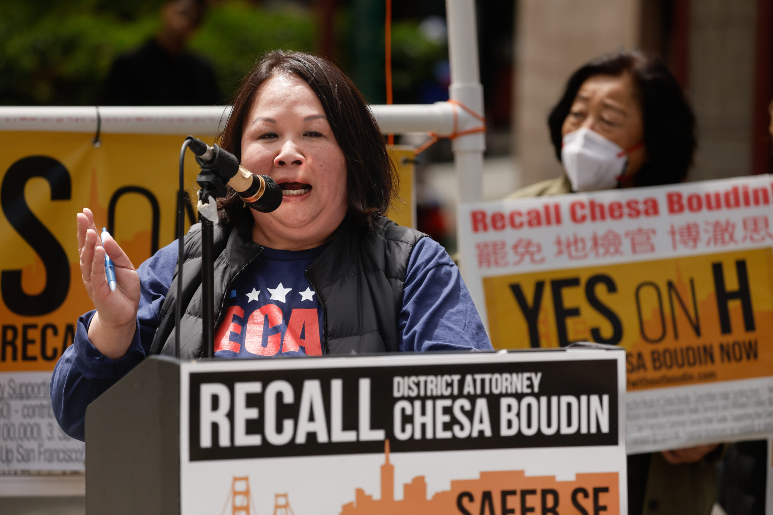Mary Jung speaks into a microphone at a podium with signs urging to recall District Attorney Chesa Boudin.