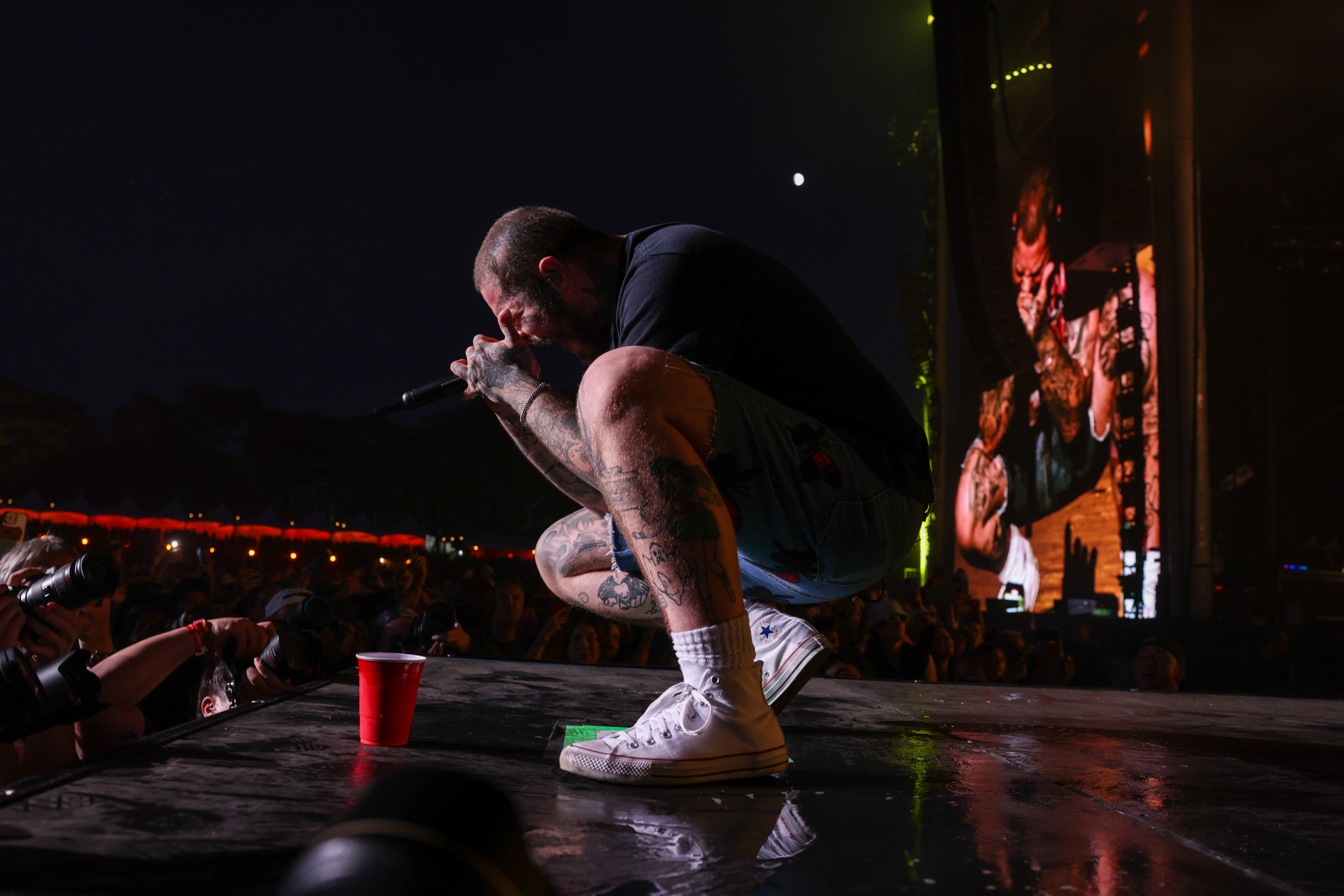 a white rapper kneels in profile on a darkened festival stage holding a microphone