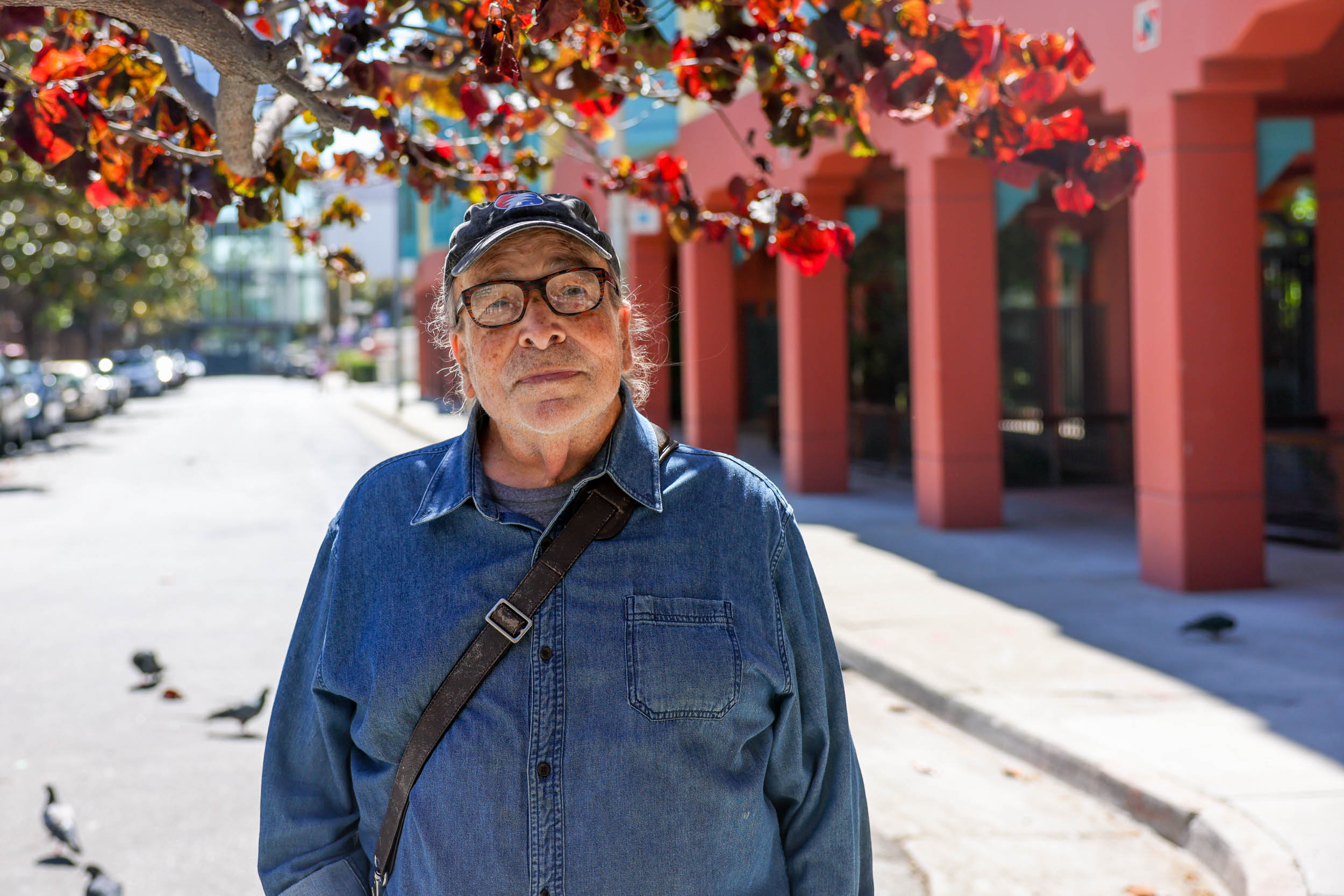 John Elberling in a denim jacket stands on a sunny street with red-leafed trees and buildings behind him.