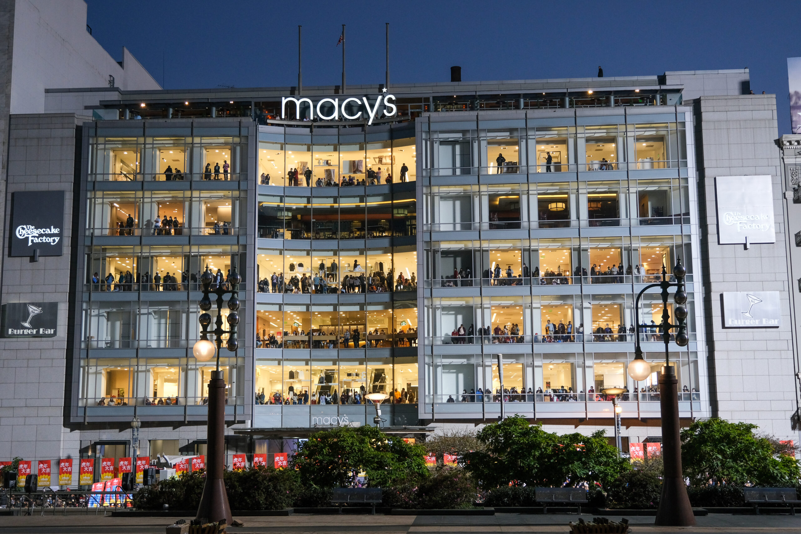 A night-time photo of a multi-story Macy's store with illuminated interiors and visible shoppers.