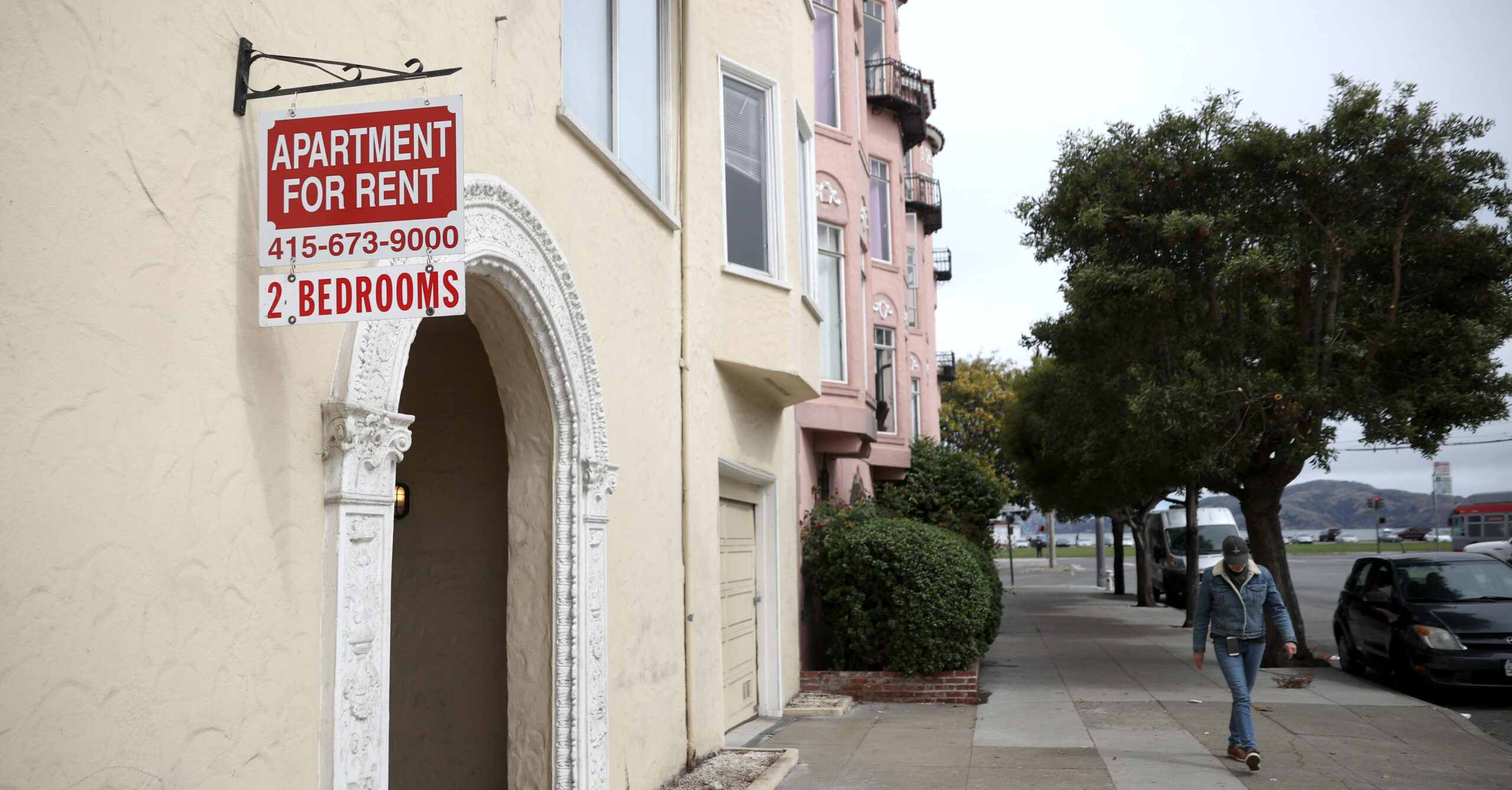 An apartment building displays a "For Rent" sign with a phone number and "2 Bedrooms." A person walks on the sidewalk beside trees and adjacent buildings.