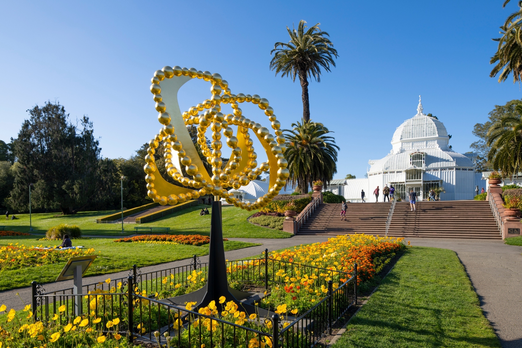 Golden spiral sculpture, colorful flowers, and a white domed conservatory under a clear blue sky.
