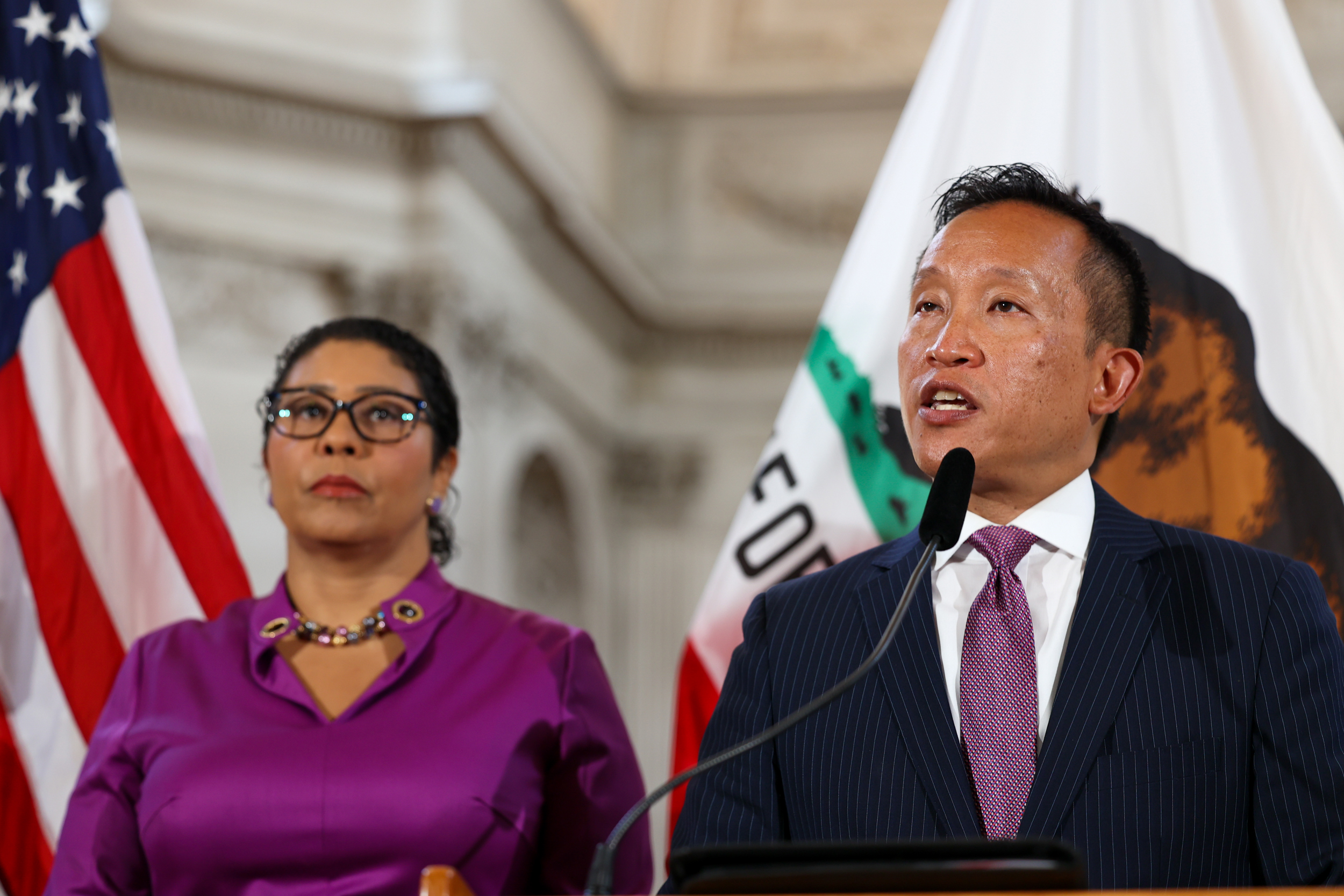 A man is speaking at a podium with microphones, a woman stands next to him, both in business attire, with the US and California flags behind them.