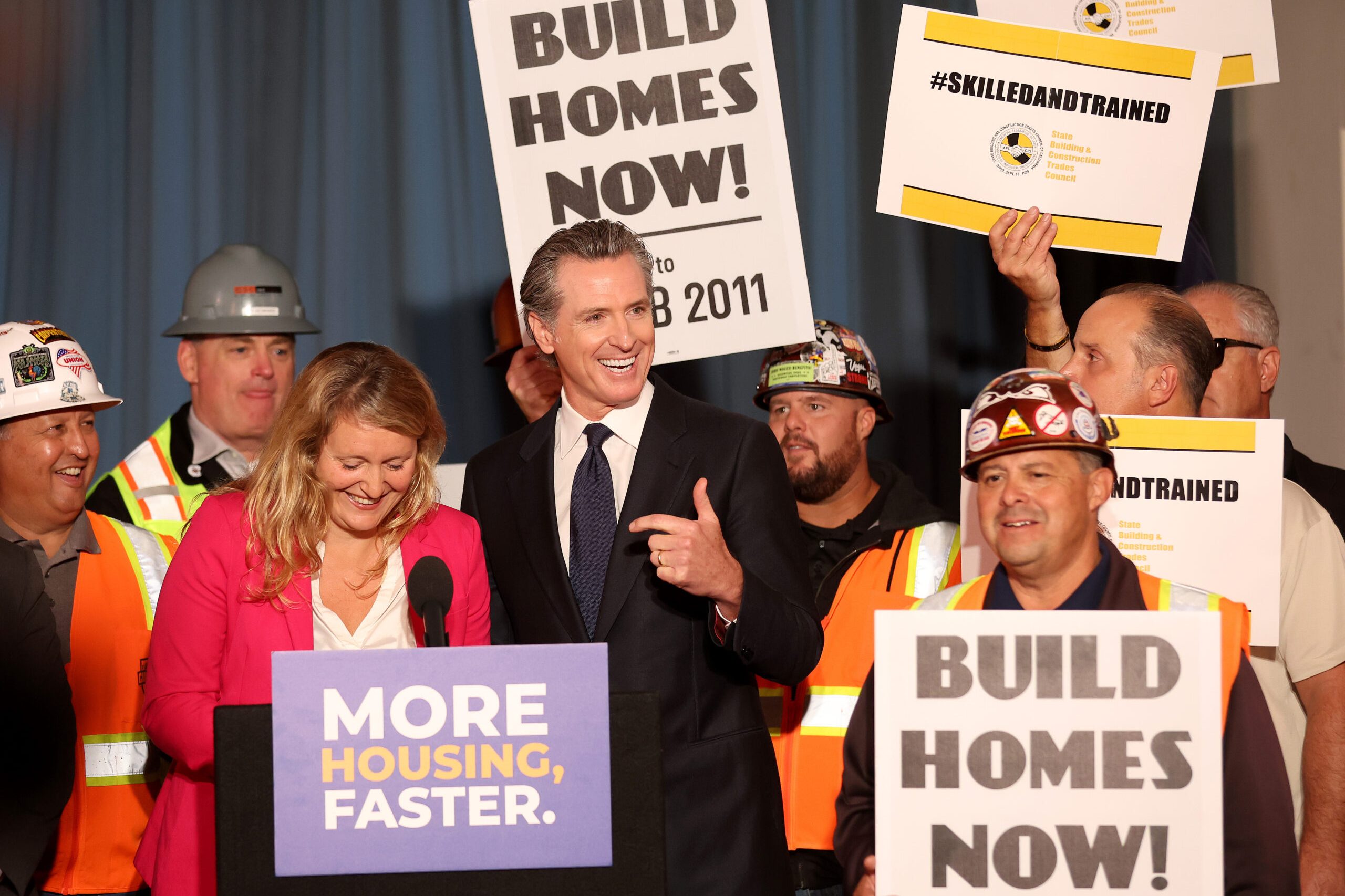 A group of people, some in construction gear, gather at a podium. Signs read "BUILD HOMES NOW!" and "MORE HOUSING, FASTER." A man in a suit speaks, smiling.