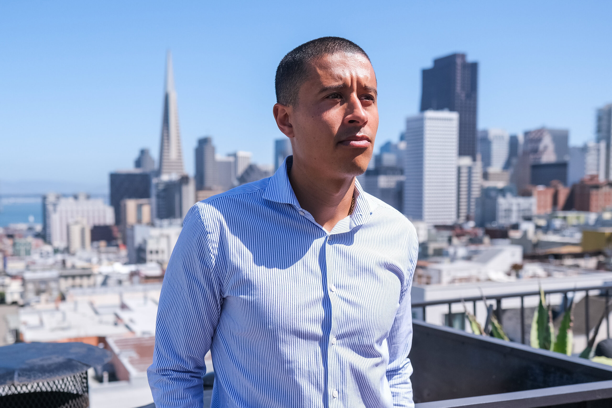 A man in a striped shirt stands on a rooftop with a city skyline in the background, featuring the Transamerica Pyramid under a clear blue sky.