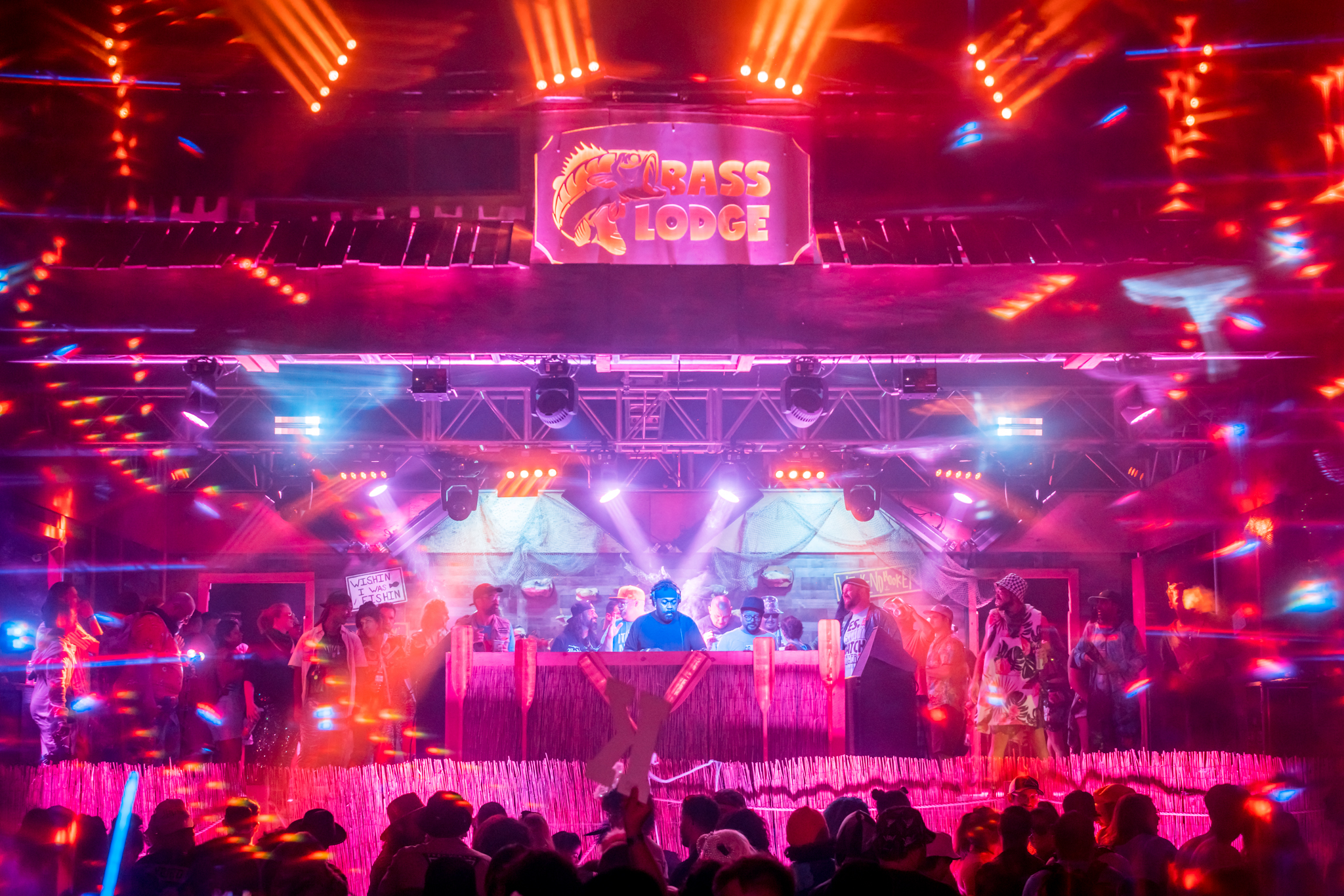 A lively music stage with vibrant multi-colored lights and a &quot;Bass Lodge&quot; sign. A DJ performs while a crowd dances below and people stand behind the DJ booth.