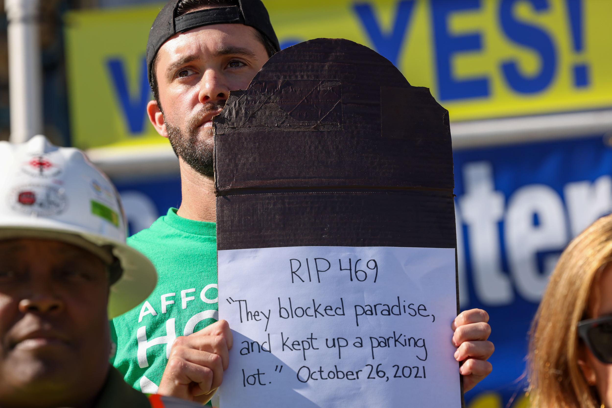 A person is holding up a handmade sign that reads "RIP 469 They blocked paradise, and kept up a parking lot." with the date "October 26, 2021".