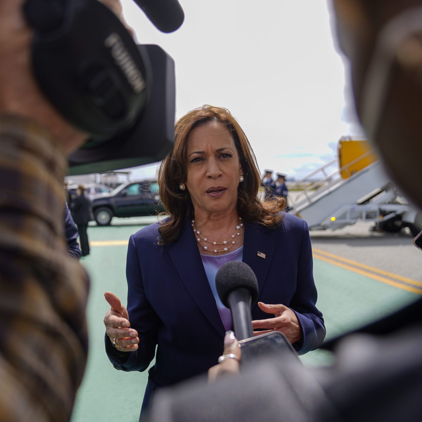 A woman in a dark suit speaks to the press outdoors, flanked by microphones and cameras. In the background, a staircase leads to an elevated area.