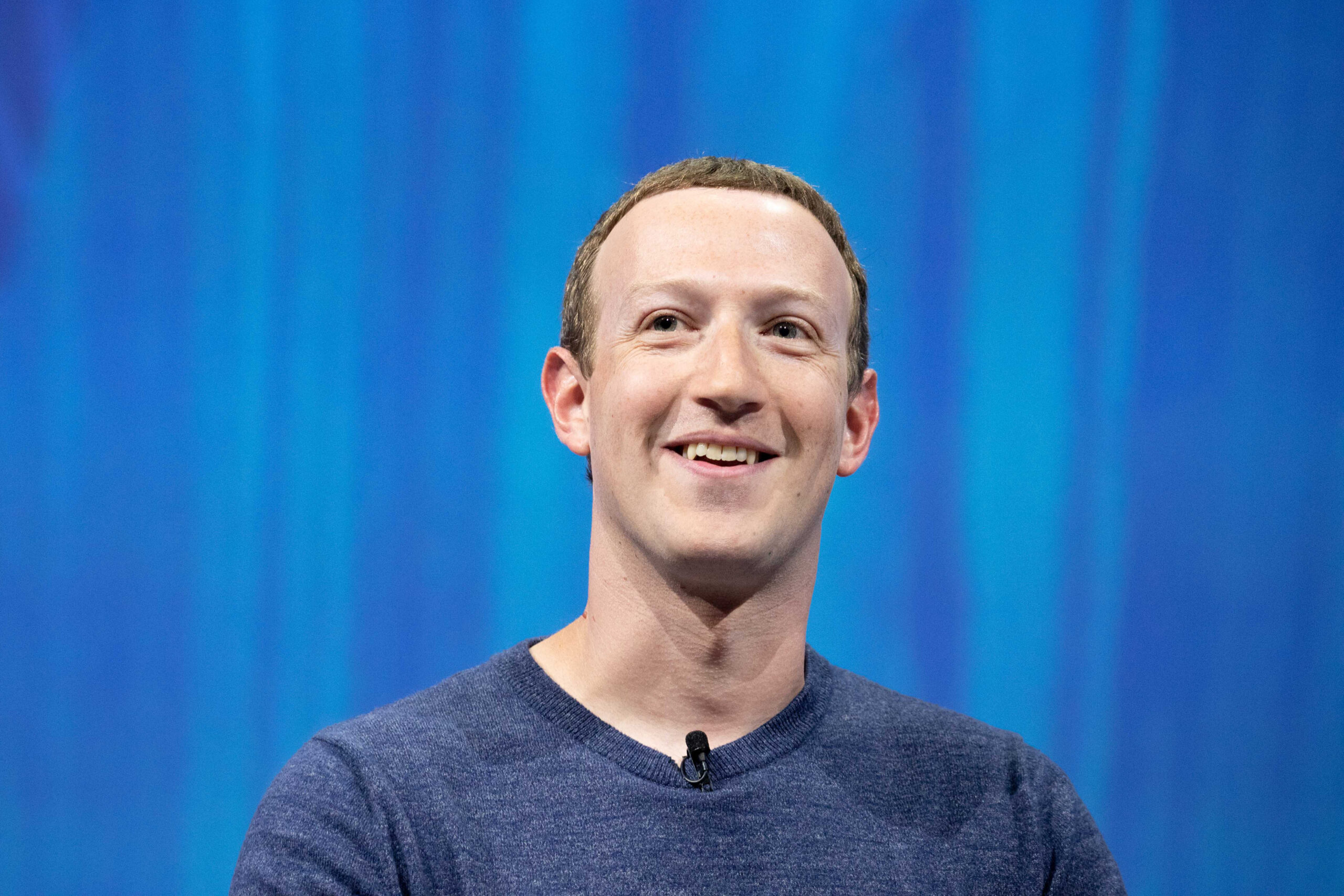 A smiling person in a blue sweater is in front of a vibrant blue background, with a black microphone clipped to their shirt.