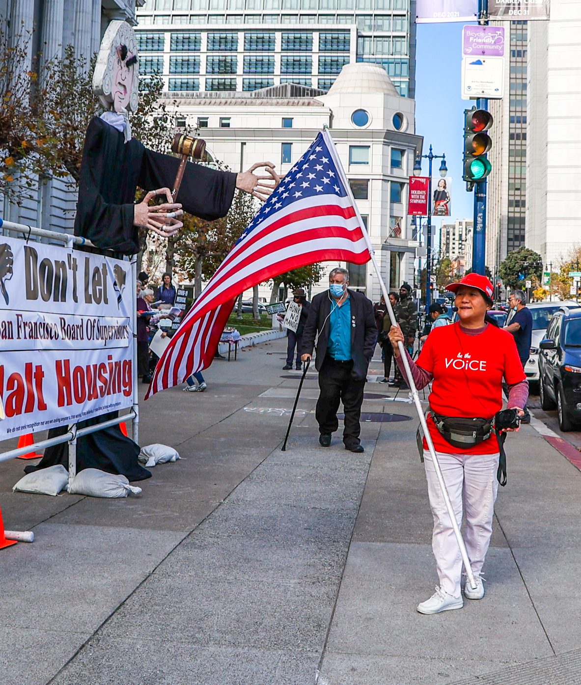 A protest scene with a person holding a large U.S. flag next to a giant puppet judge, and a banner about housing.