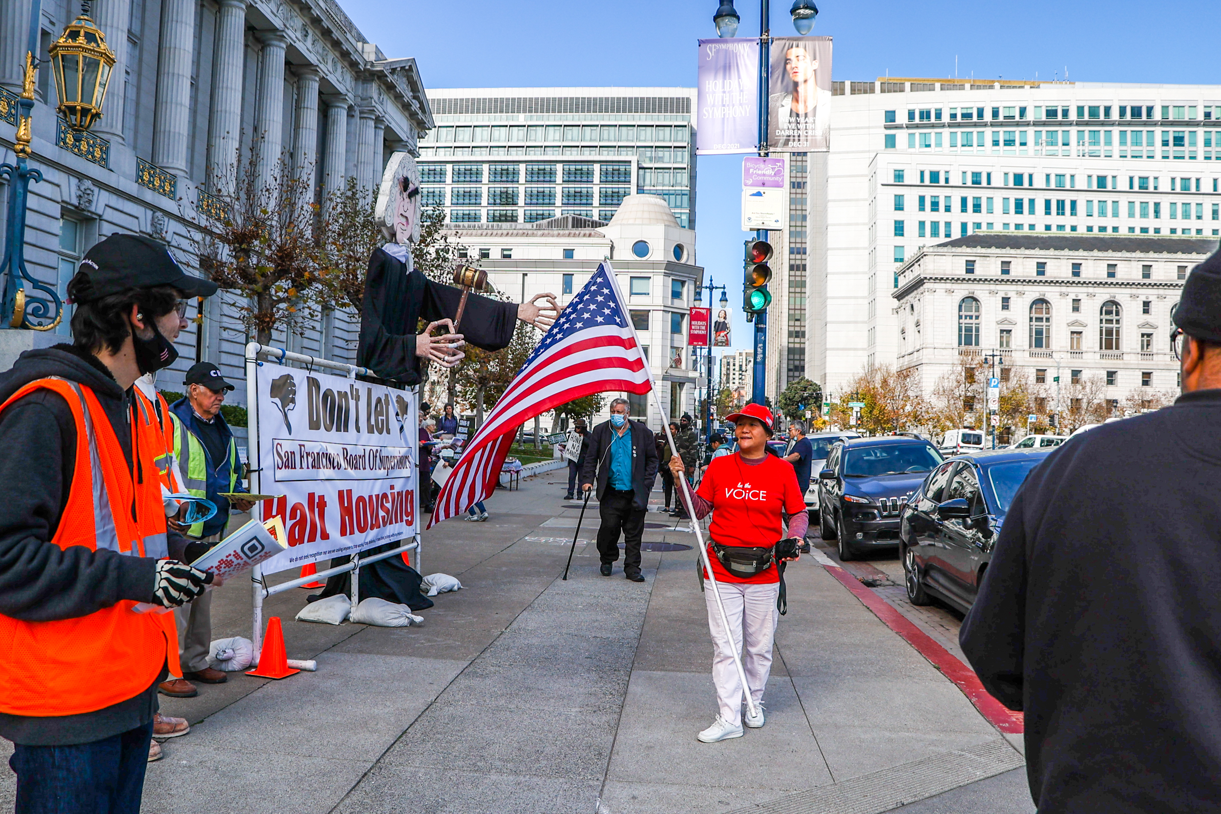 A protest scene with a person holding a large U.S. flag next to a giant puppet judge, and a banner about housing.