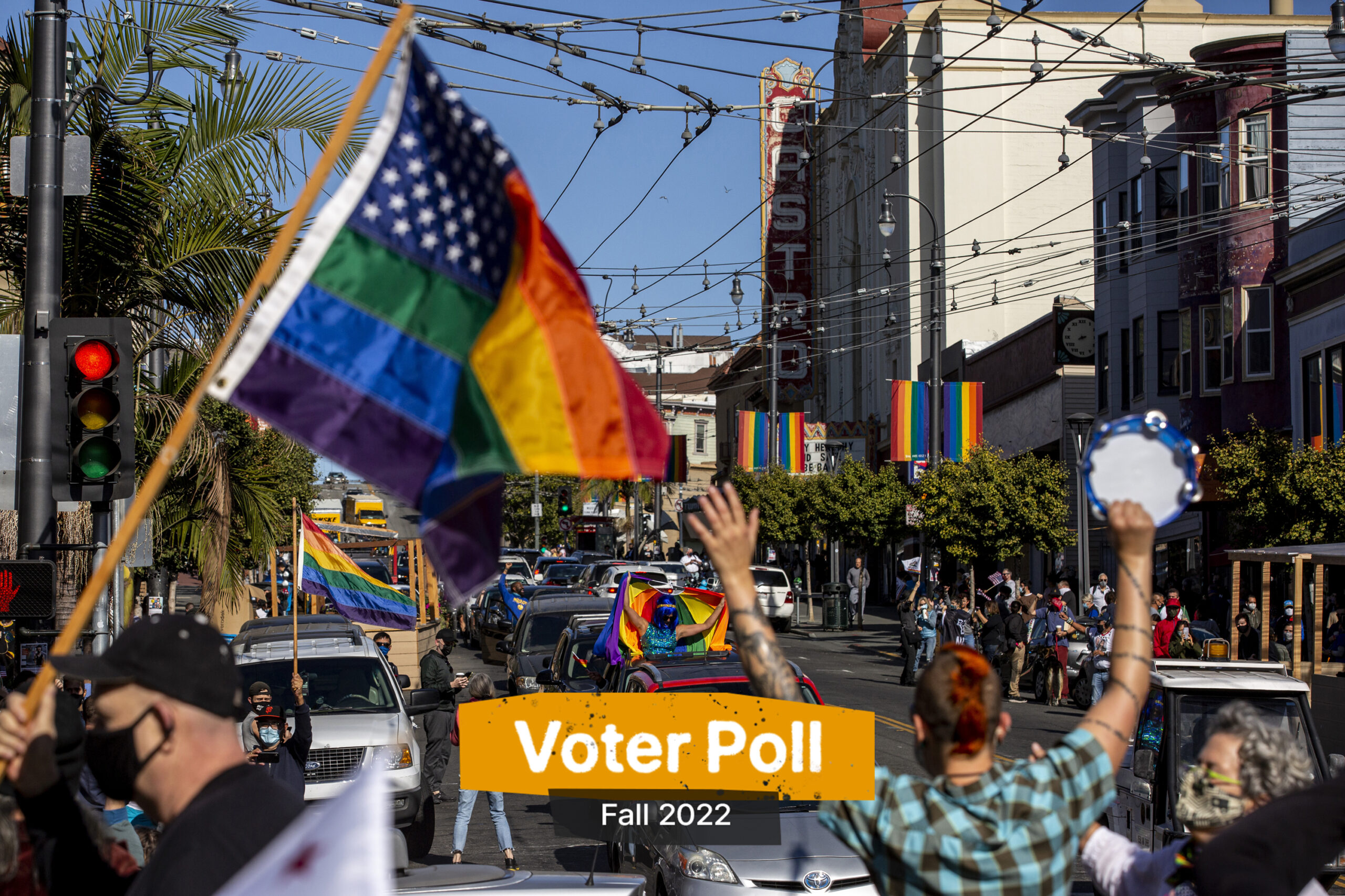 Image of Castro Street, with a gay pride rainbow/american flag flying on the left side. A person with a red mohawk holds a tambourine up as they look down the street towards cars.