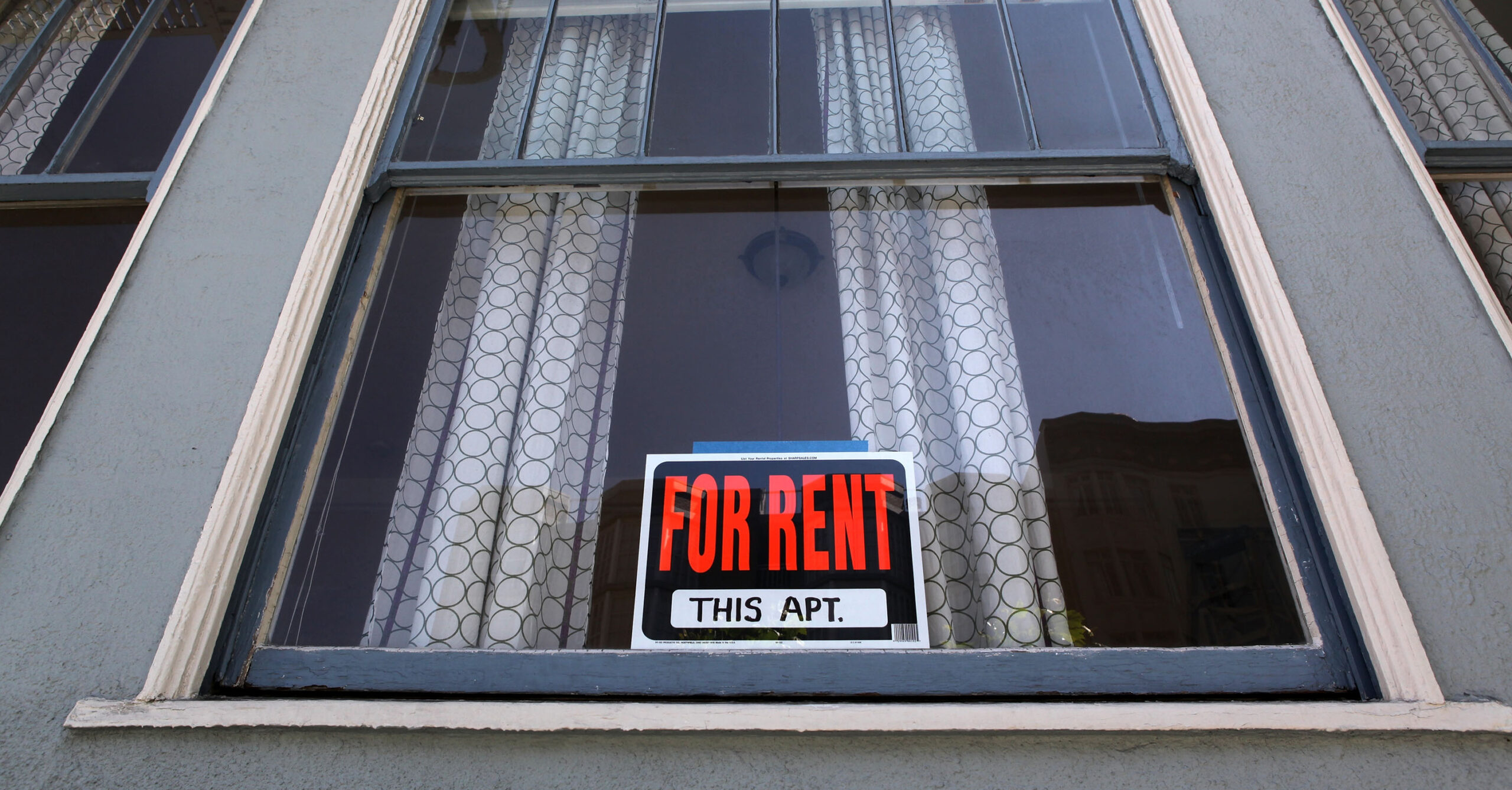 A window with a "FOR RENT" sign is displayed. The sign is in red and black lettering, set against curtains with a circular pattern. The building has a gray exterior.