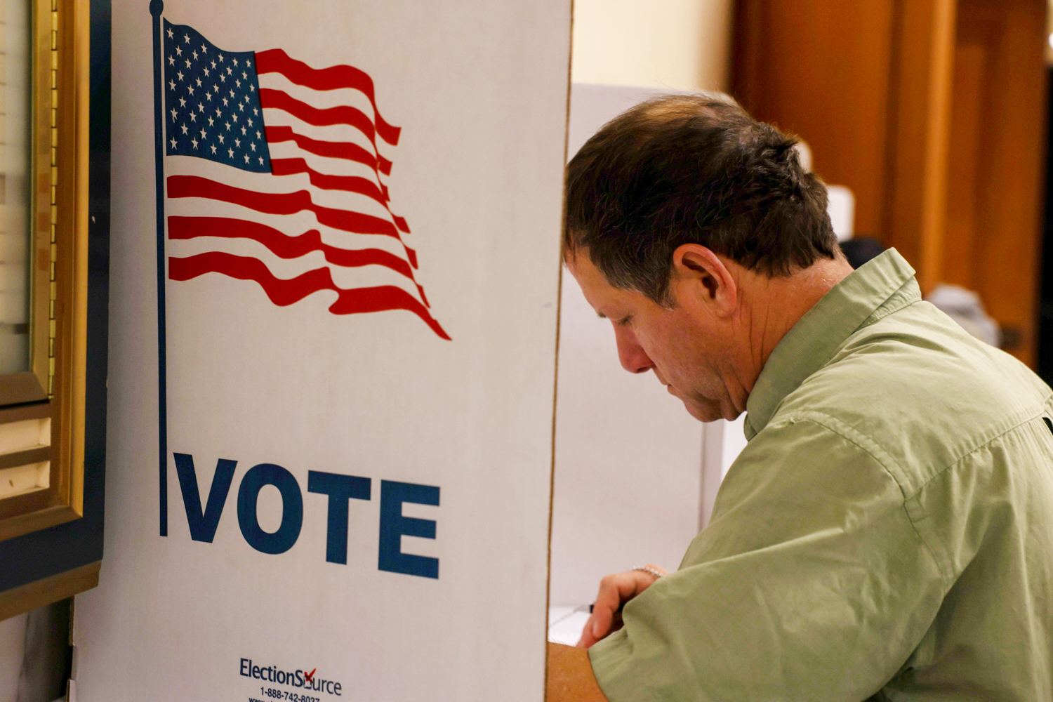 A man is filling out a ballot at a voting booth marked with an American flag and "VOTE."