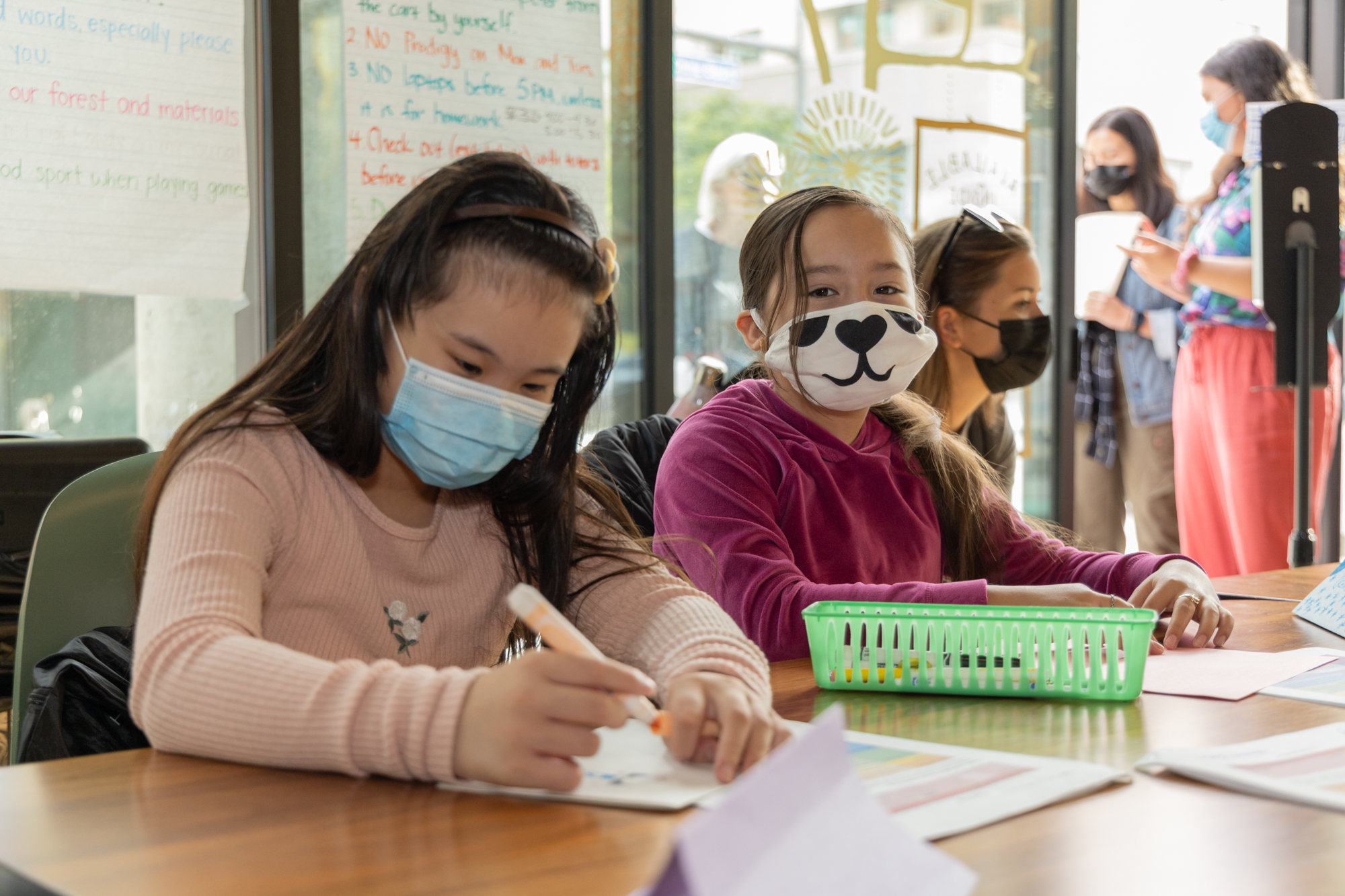 Two girls sit at a desk wearing masks, one has a mask with a cat design, engaging in a classroom activity.