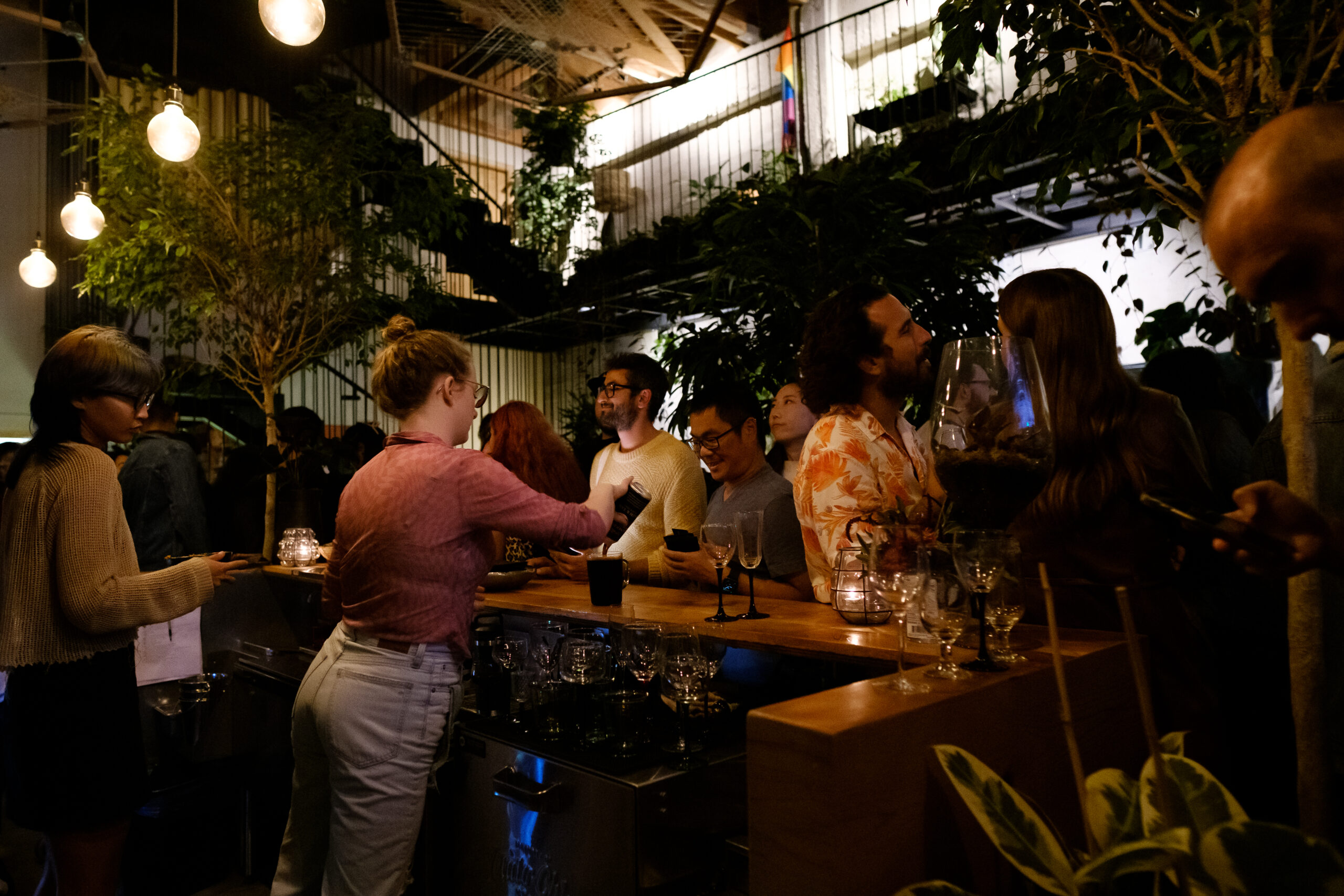 The image shows a lively bar scene with people chatting and drinking under warm lighting. Plants and hanging bulbs decorate the cozy, bustling space.