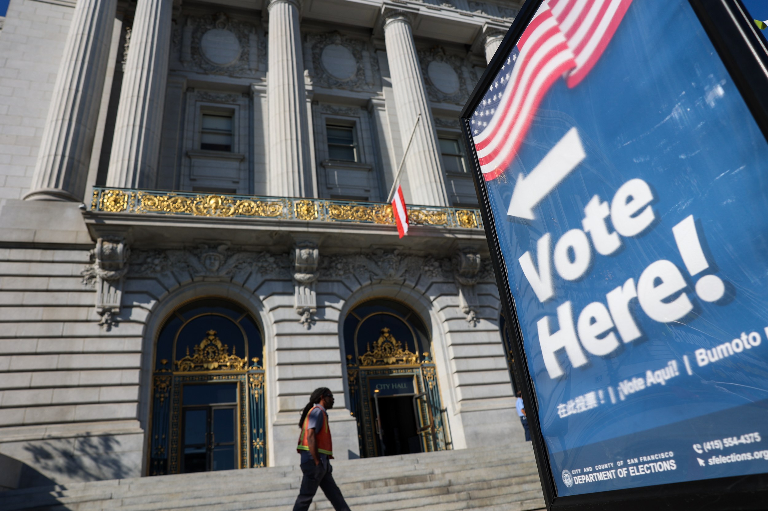 A person walks by a grand building with a "Vote Here!" sign in the foreground.