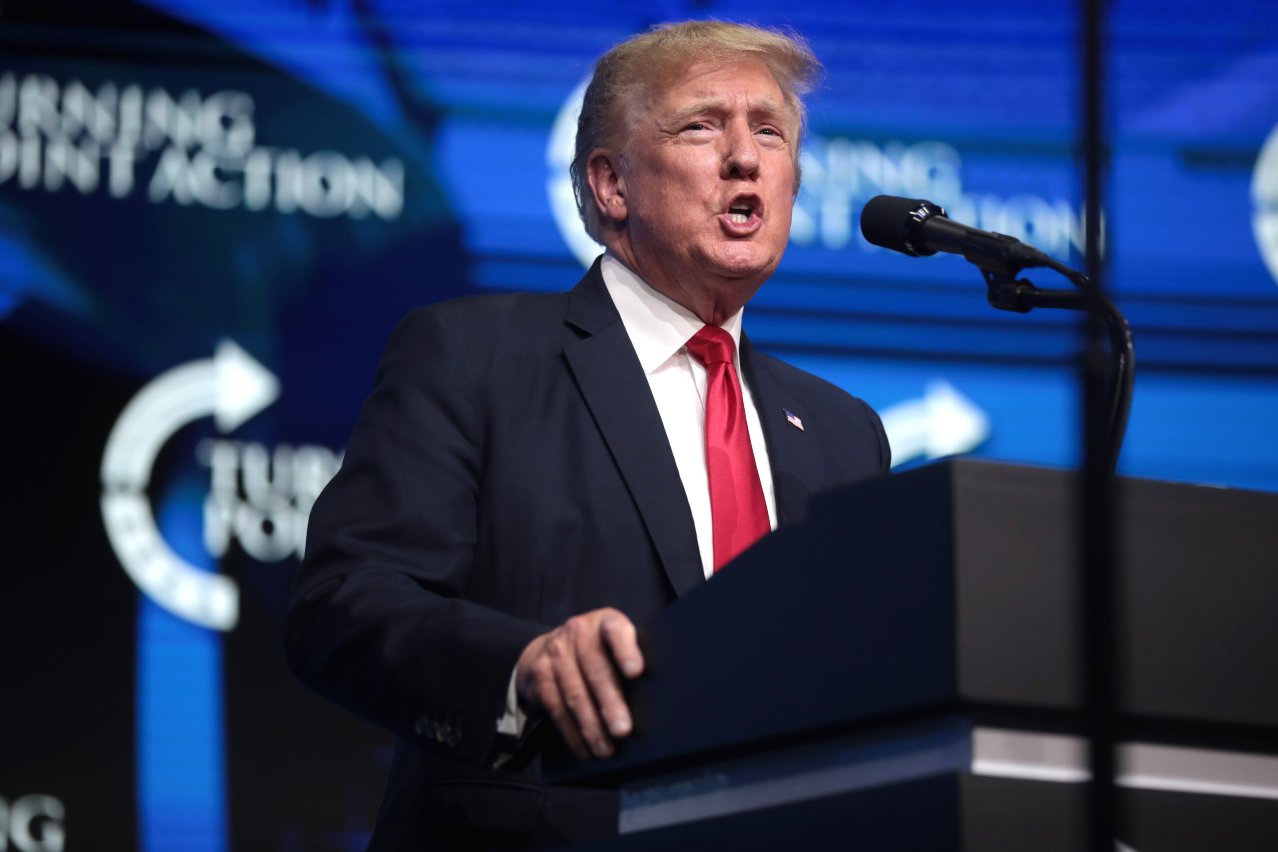 A man in a dark suit with a red tie speaks passionately at a podium with a microphone against a blue backdrop featuring the words "Turning Point Action."