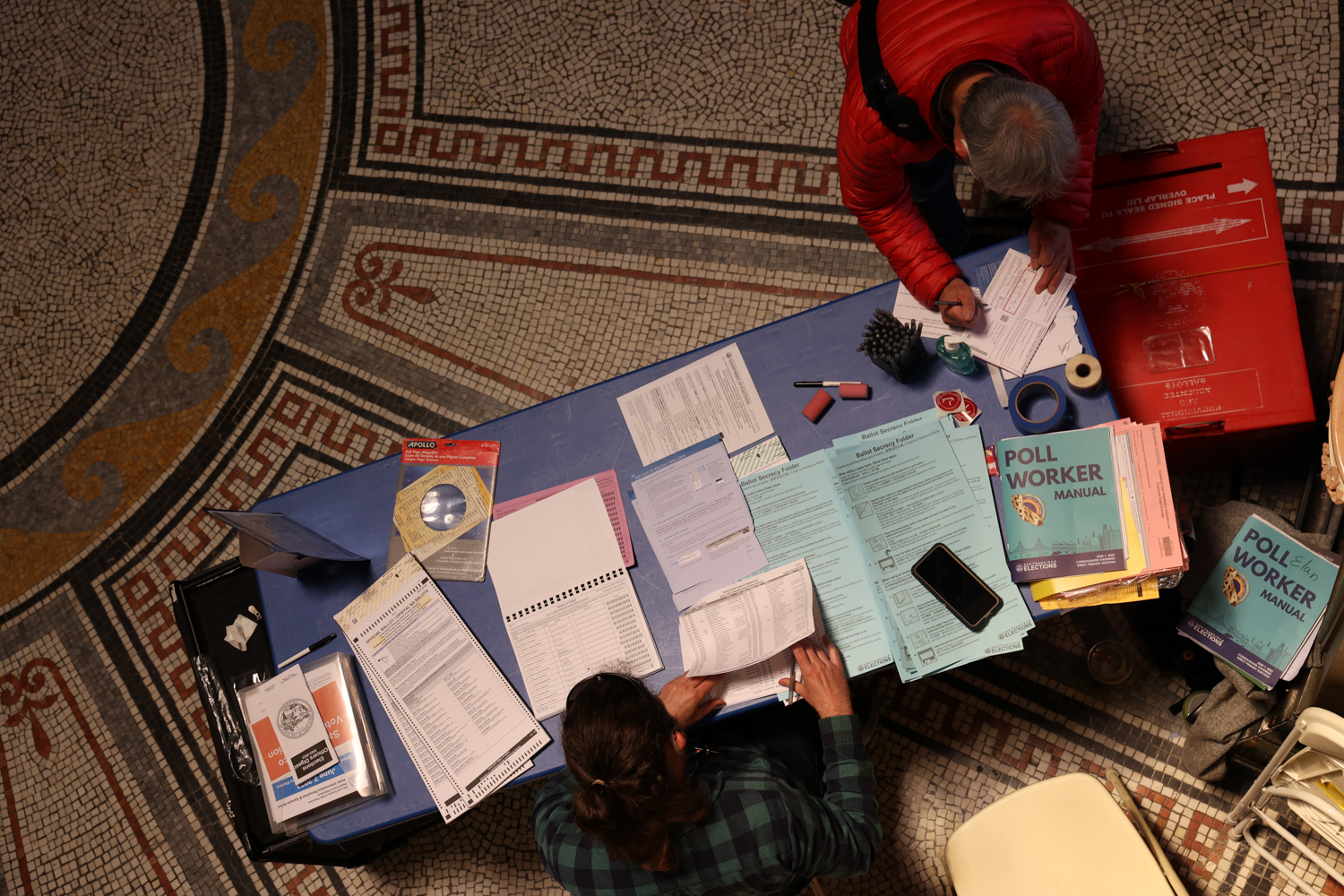 Two individuals are at a table with paperwork, possibly in a voting area, with a decorative tile floor visible.