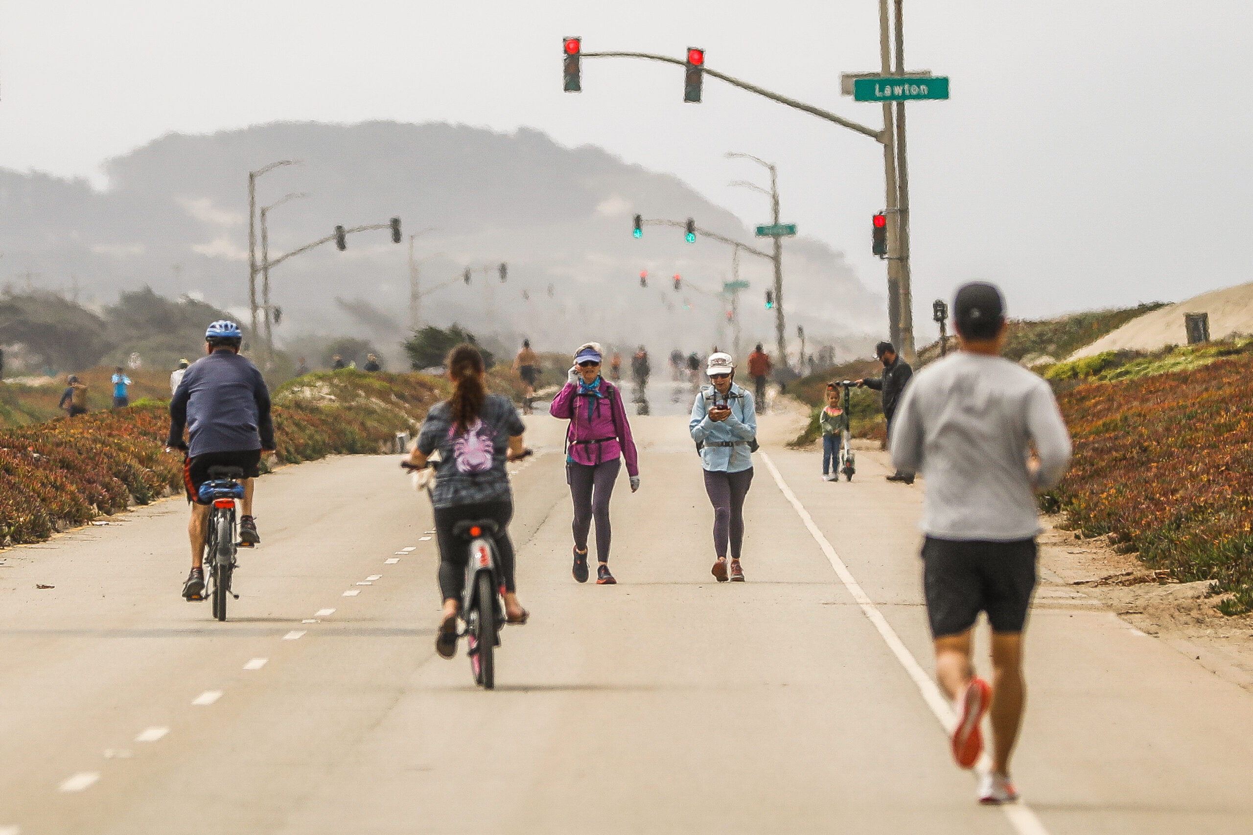 A road with cyclists, walkers, and a runner, with traffic lights overhead and foggy hills in the distance.