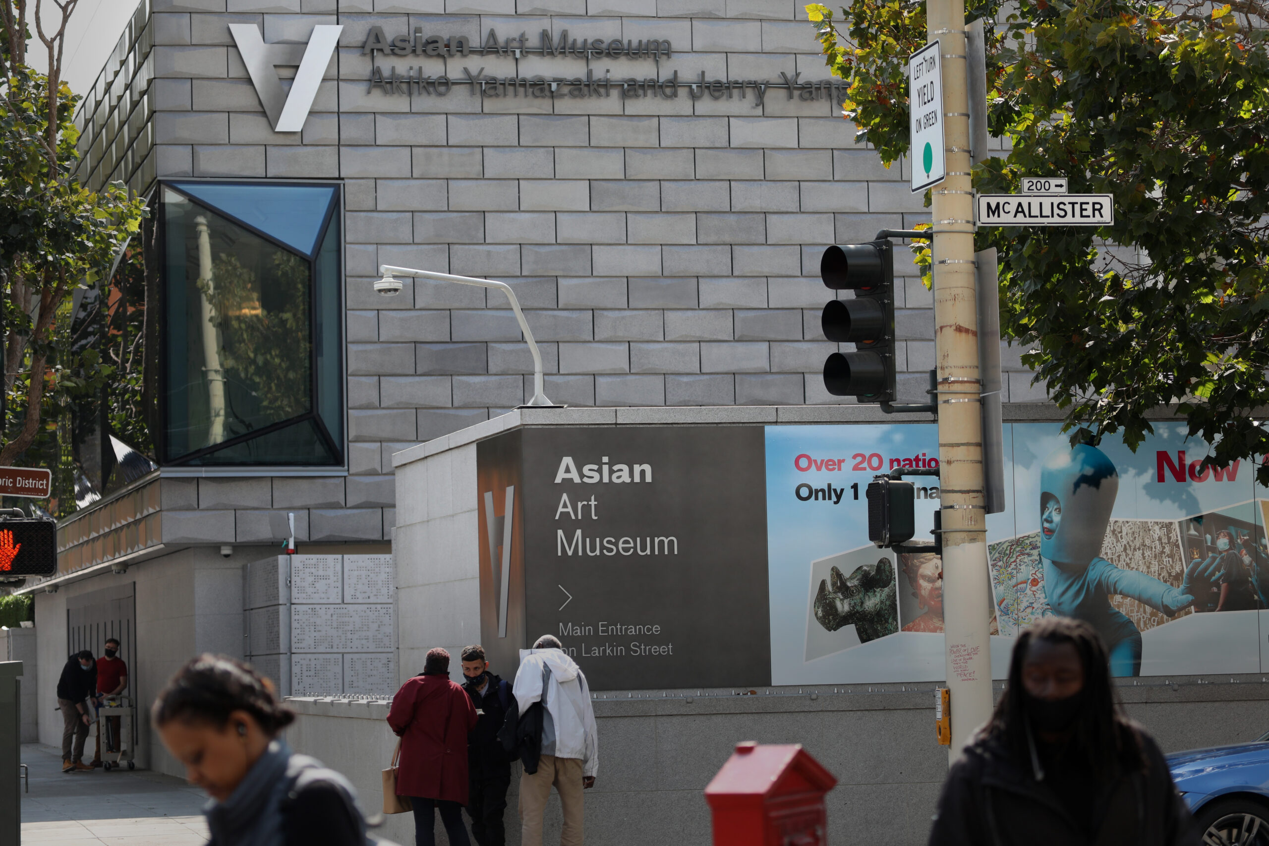 The image shows the exterior of the Asian Art Museum with a modern facade, street signs for McAllister and traffic lights, and people walking past.