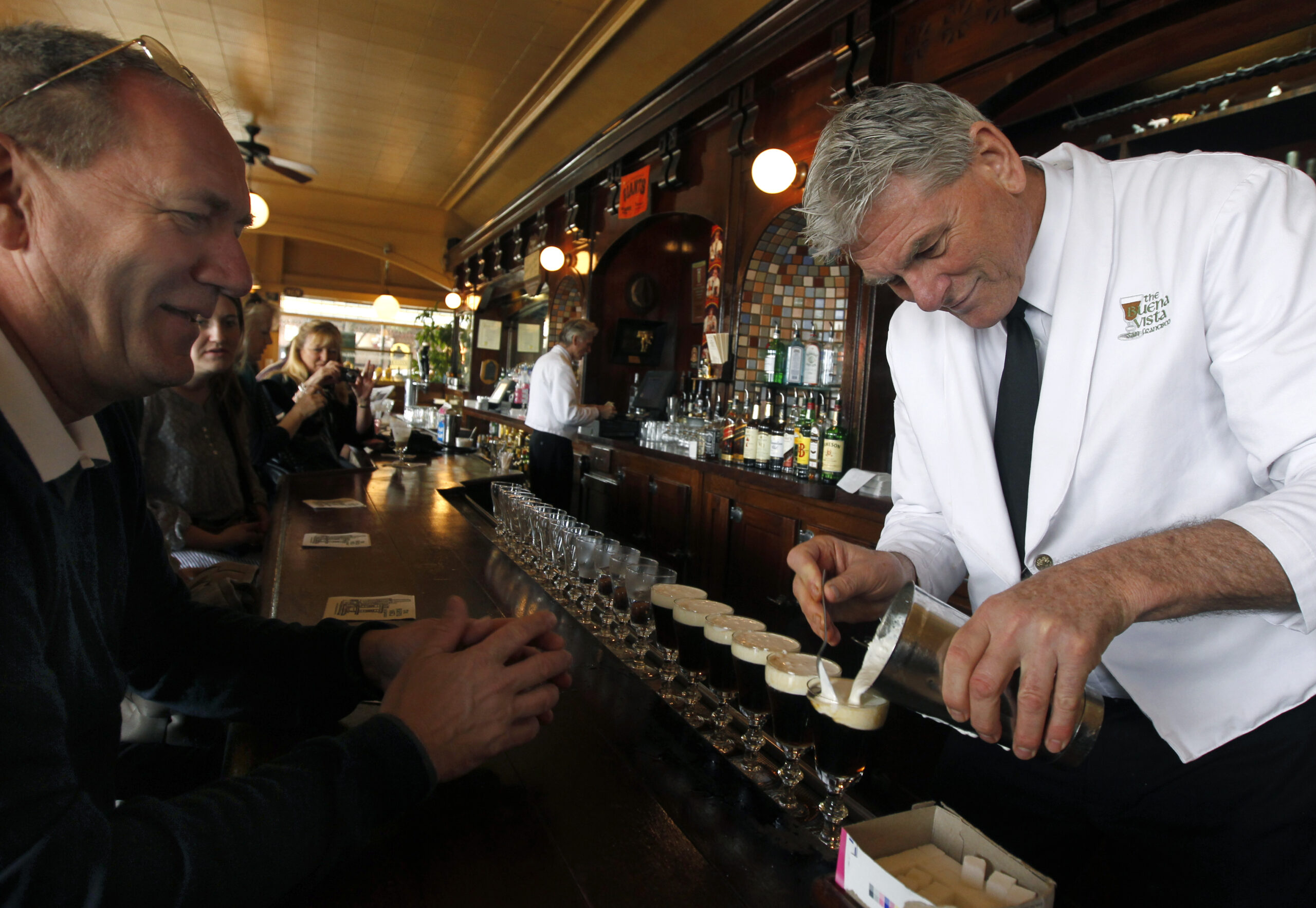 A man pours coffee into a line of clear glasses on a bar.