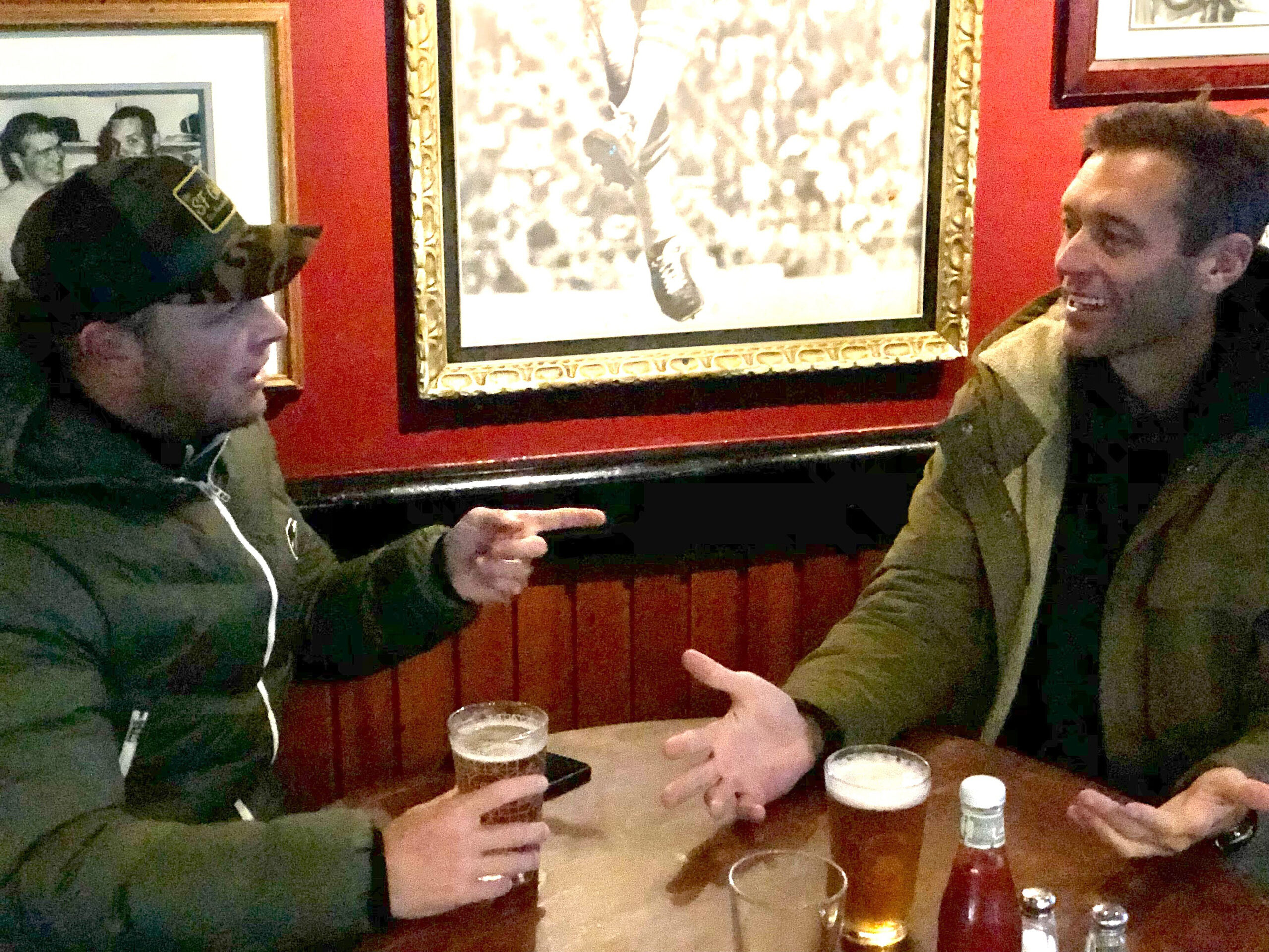 Two men in jackets sit at a wooden table in a cozy pub, talking animatedly. Each has a pint of beer, with framed pictures on the red walls behind them.