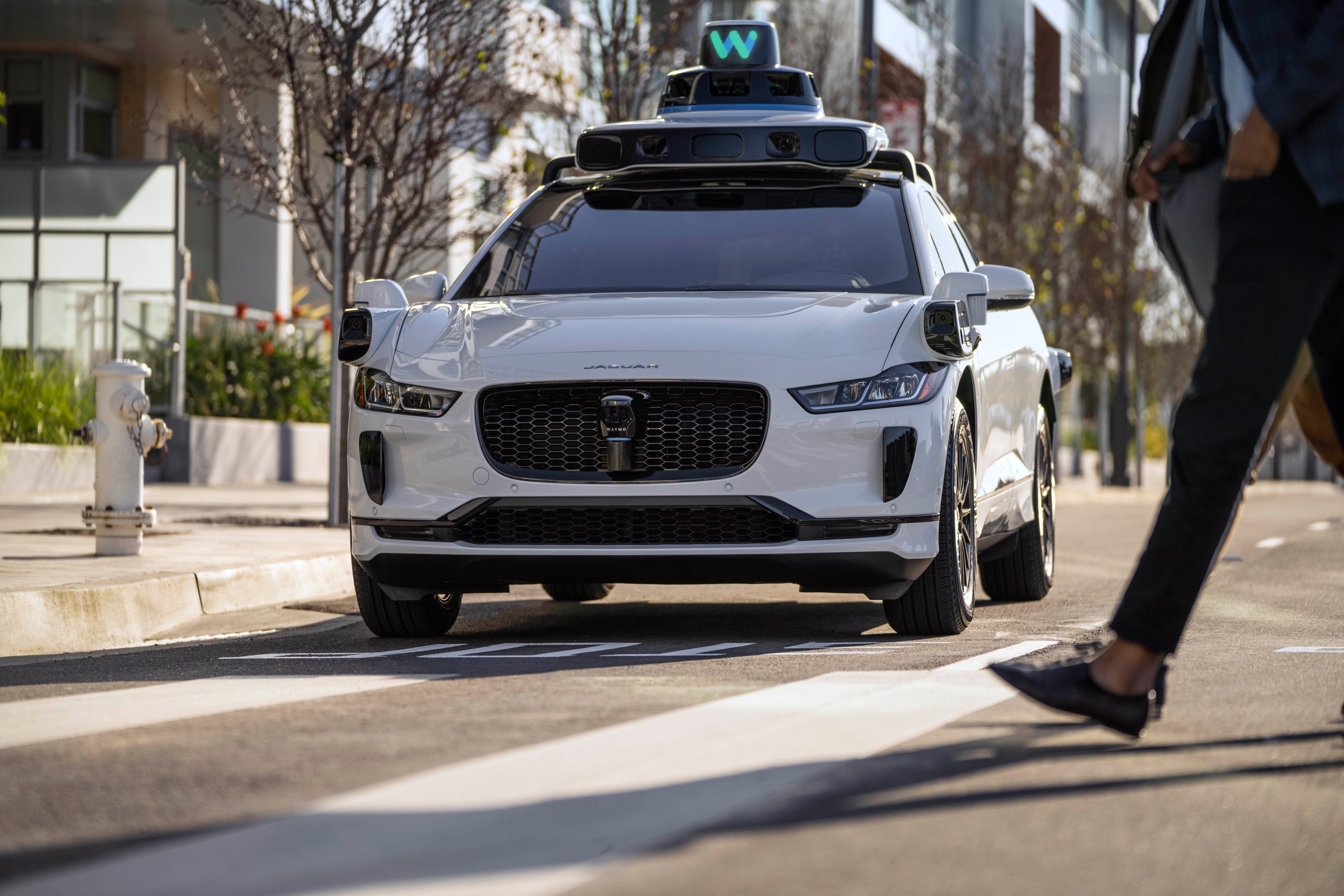 A white self-driving car with sensors on top stops at a crosswalk as a pedestrian walks by. It's on a city street lined with modern buildings and trees.